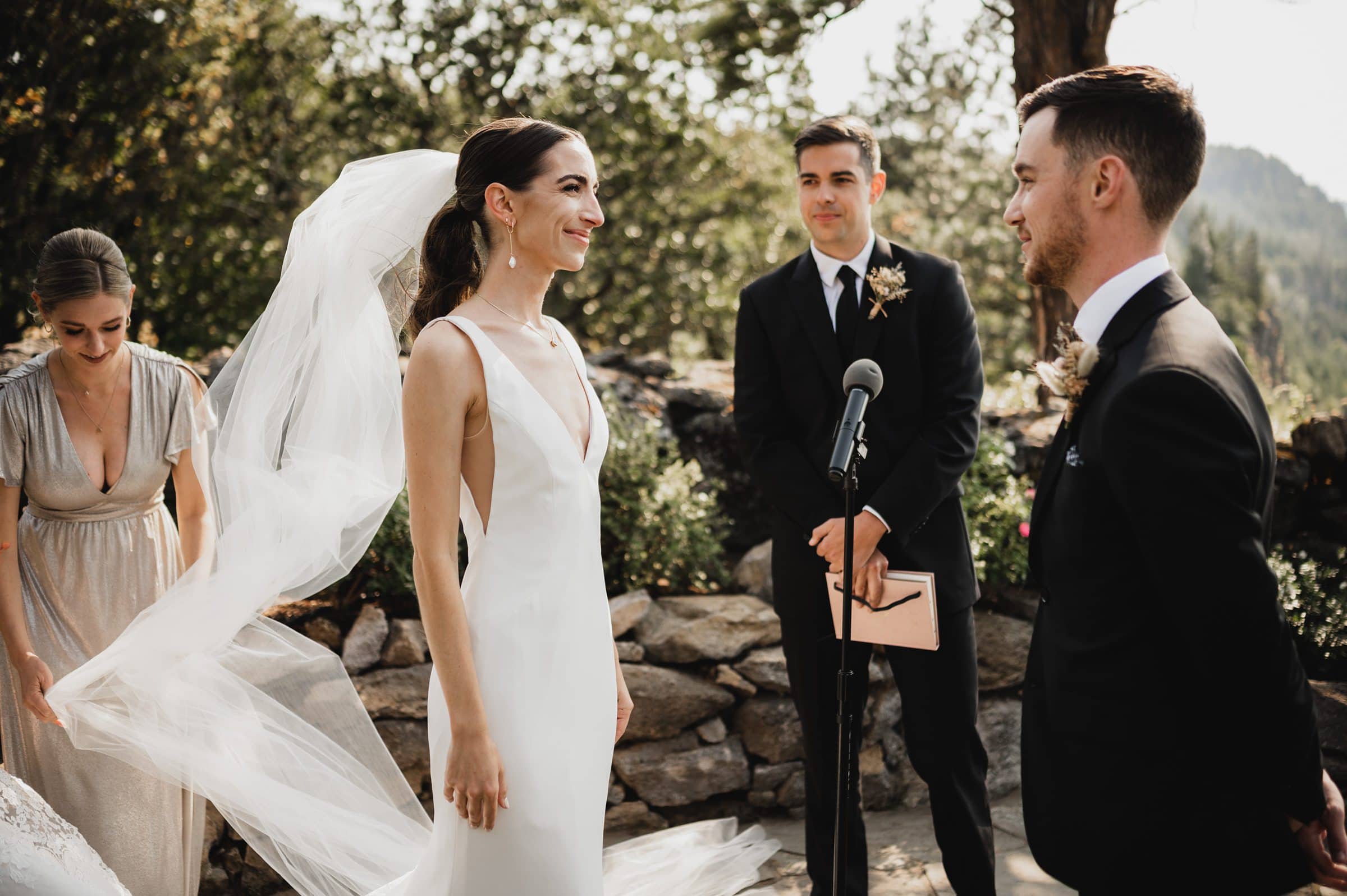 The bride and groom exchange vows outdoors at The Griffin House, as the bridesmaid adjusts the brides veil and the officiant holds a book.