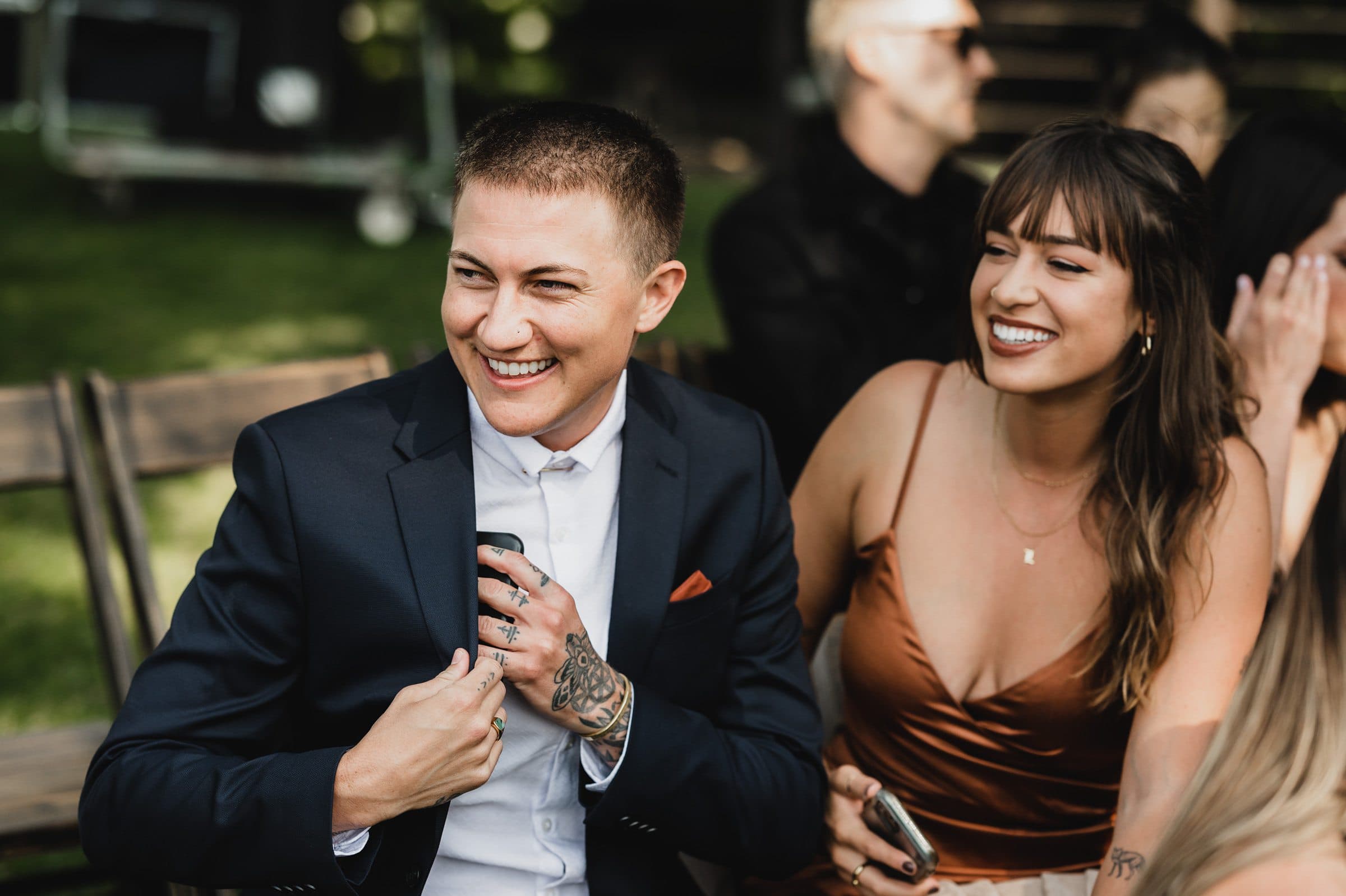 Two people sitting outdoors, smiling, one in a suit and the other in a brown dress, holding phones amidst the picturesque backdrop of The Griffin House.
