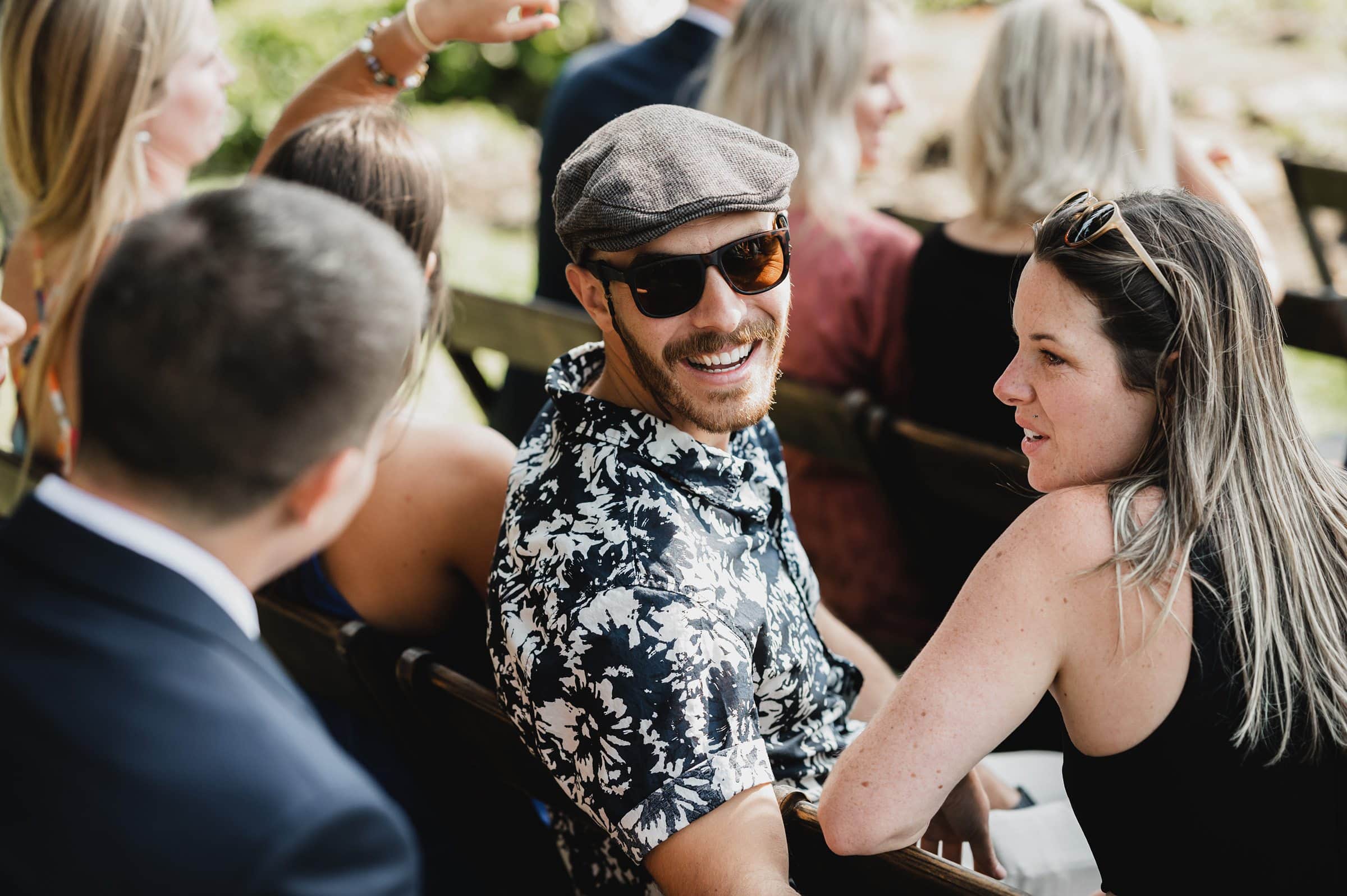 Man in sunglasses and cap smiling, seated outdoors with a group of people in casual clothing, capturing joyful moments like wedding photos at The Griffin House.