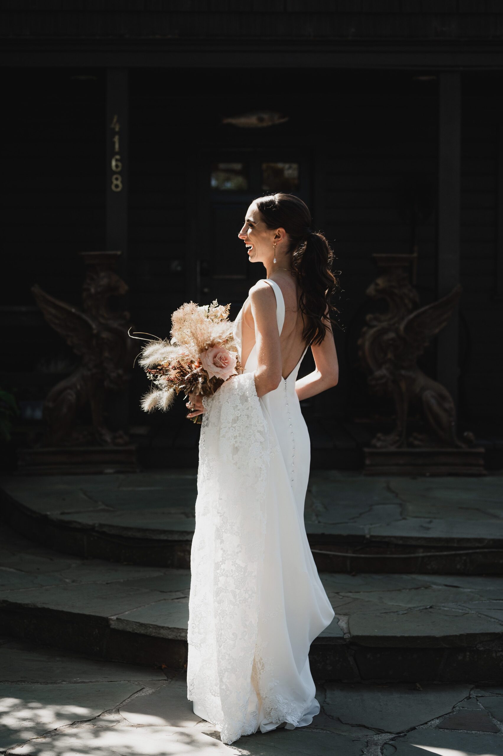 The bride in a white dress holding a bouquet stands on a stone path, with the dark backdrop of The Griffin House enhancing her wedding photos.