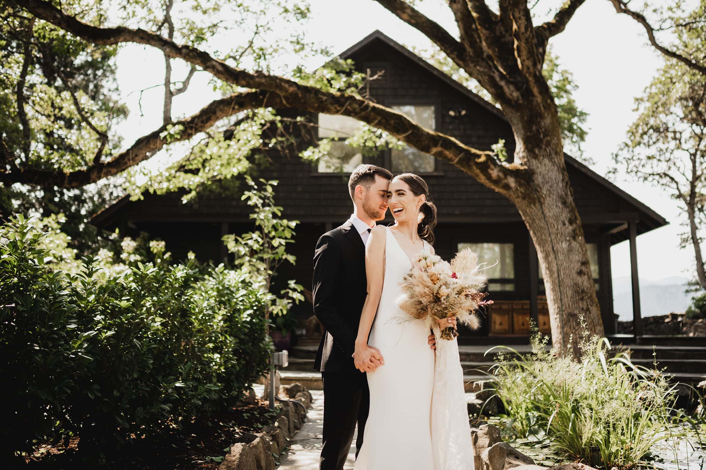 The bride and groom stand smiling in front of a tree and a house at The Griffin House, holding hands and a bouquet of pampas grass.