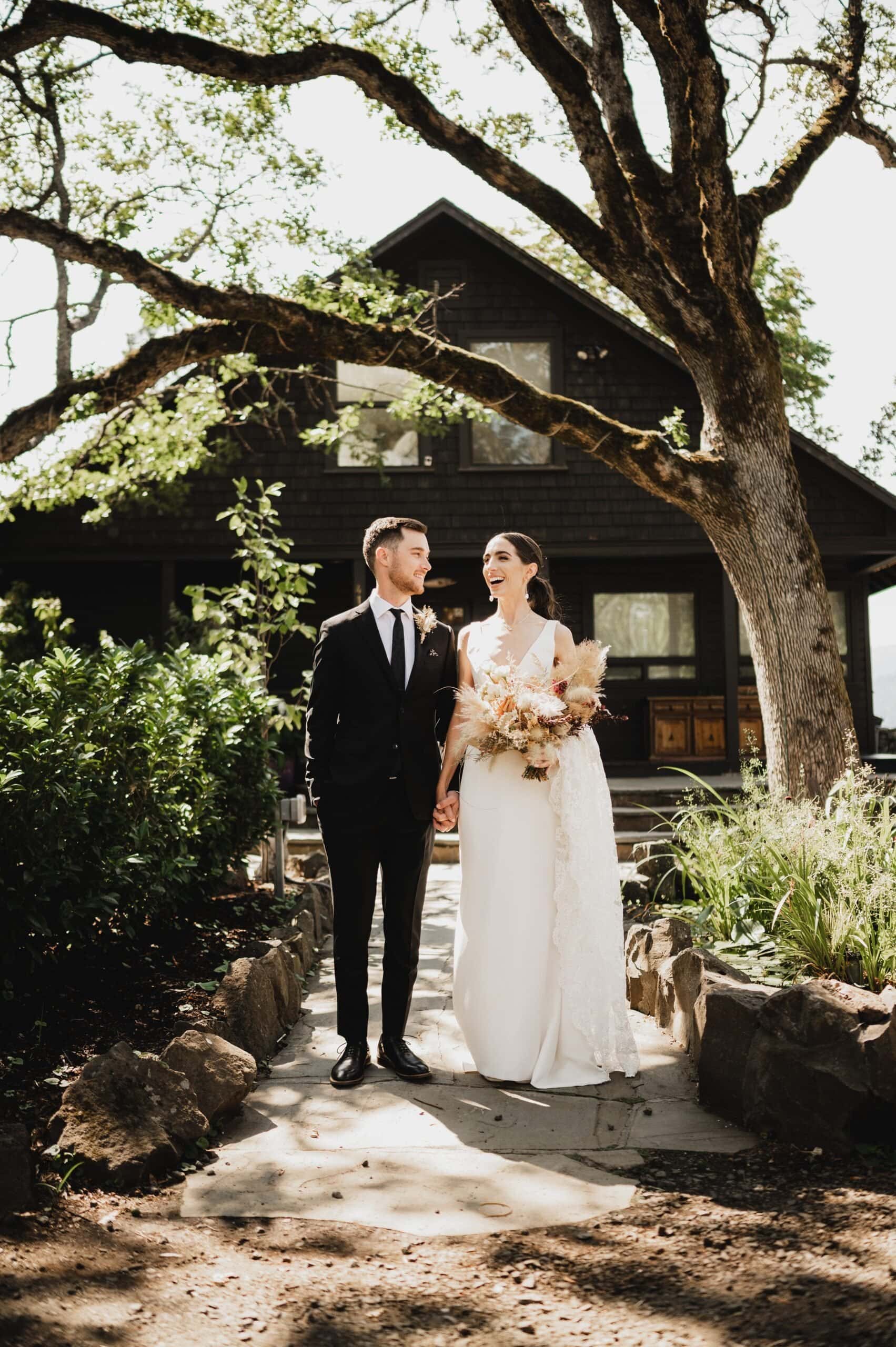 The bride and groom, hand in hand, stroll along a garden path with a charming house and tree in the background, creating perfect wedding photos at The Griffin House.