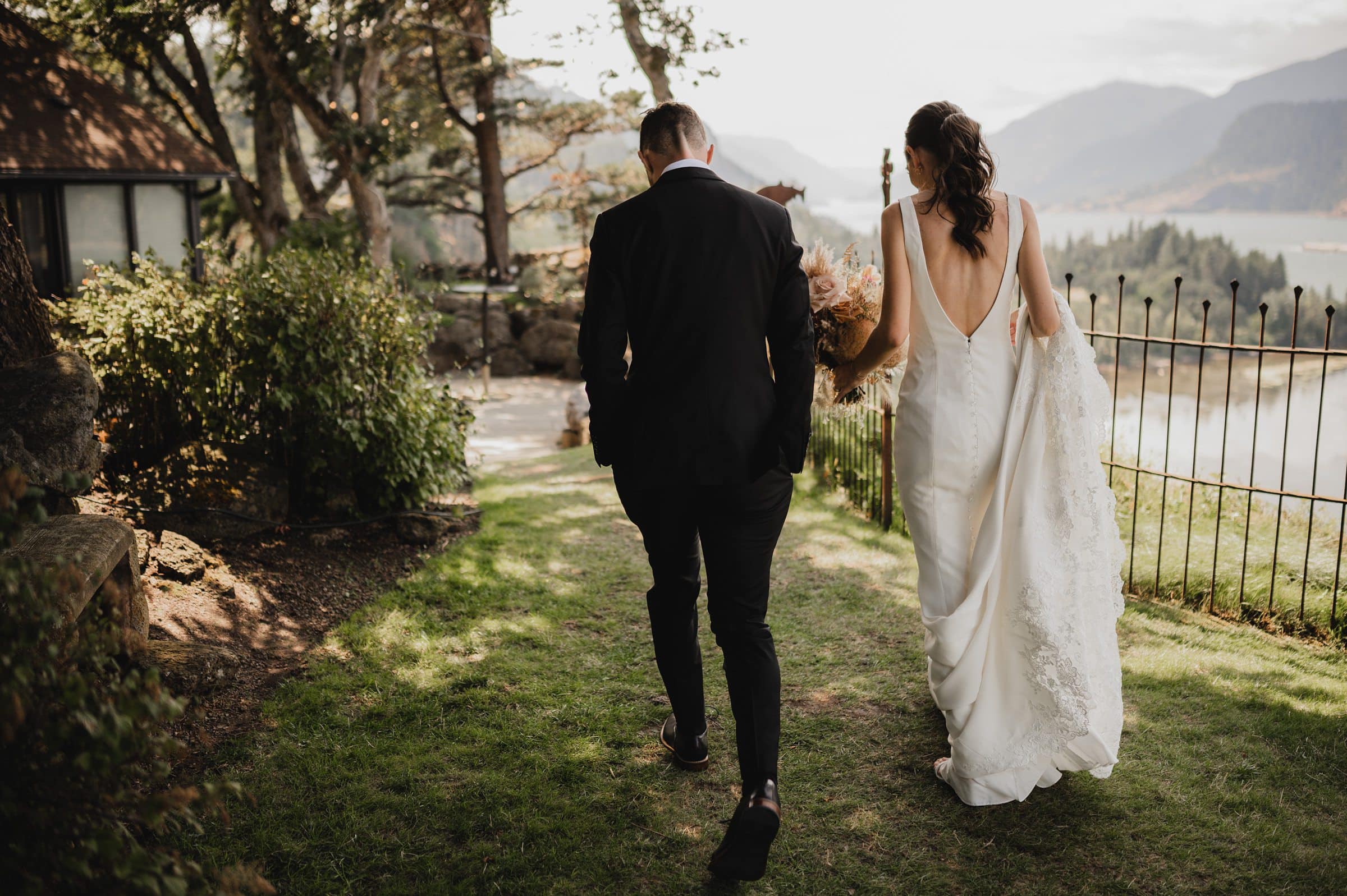 Bride and groom walk hand in hand down a grassy path with scenic mountain and lake views in the background, capturing perfect wedding photos at The Griffin House.