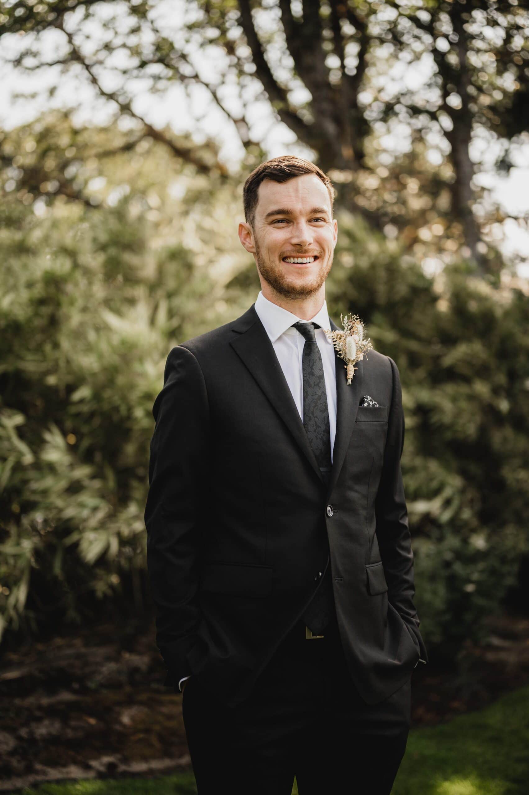 A man in a black suit with a floral boutonniere smiles while standing outdoors, surrounded by trees. The scene captures the joy of wedding photos at The Griffin House.