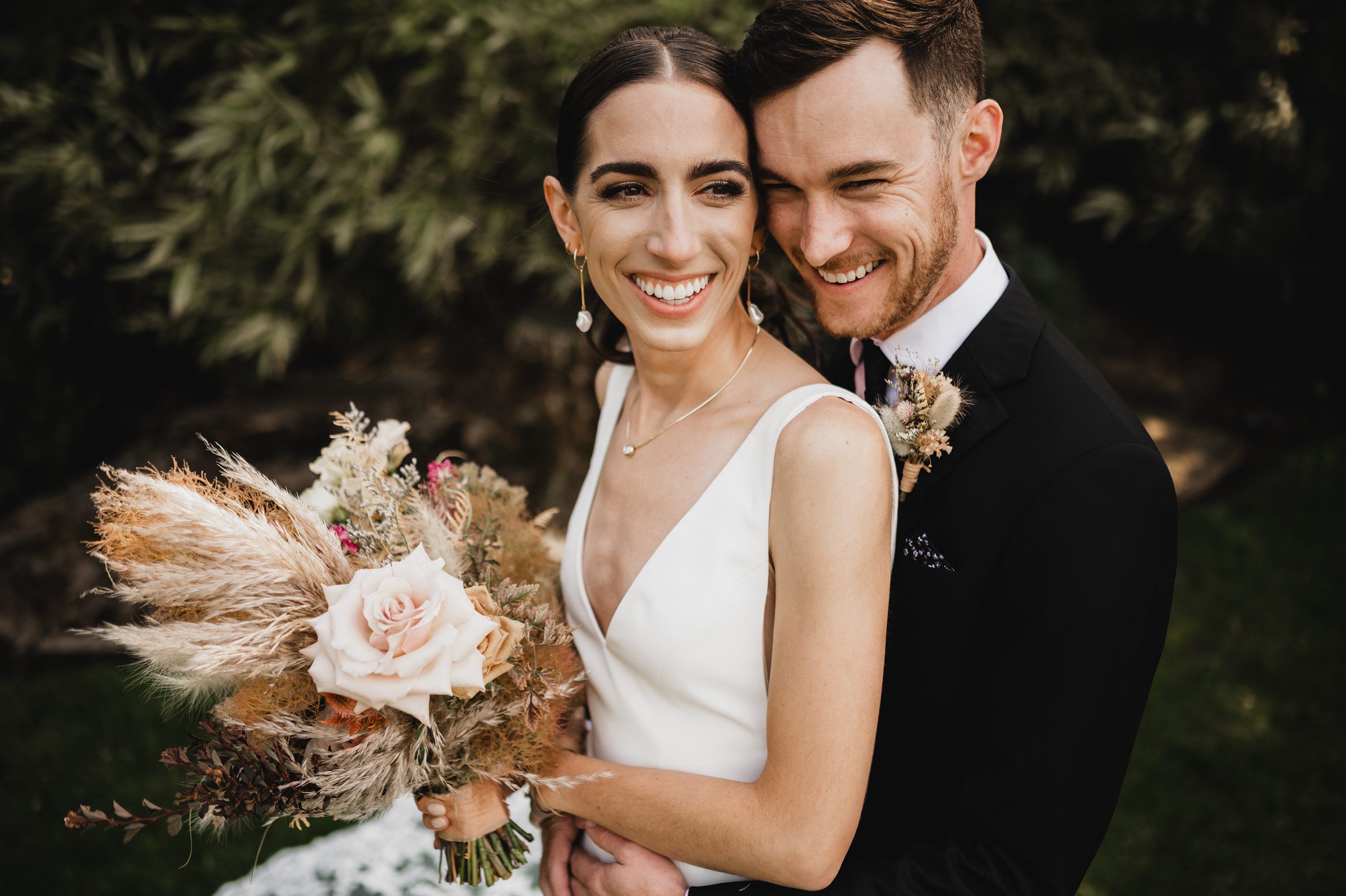The bride and groom smile warmly as they embrace outdoors at The Griffin House, with the bride elegantly holding a bouquet of flowers.