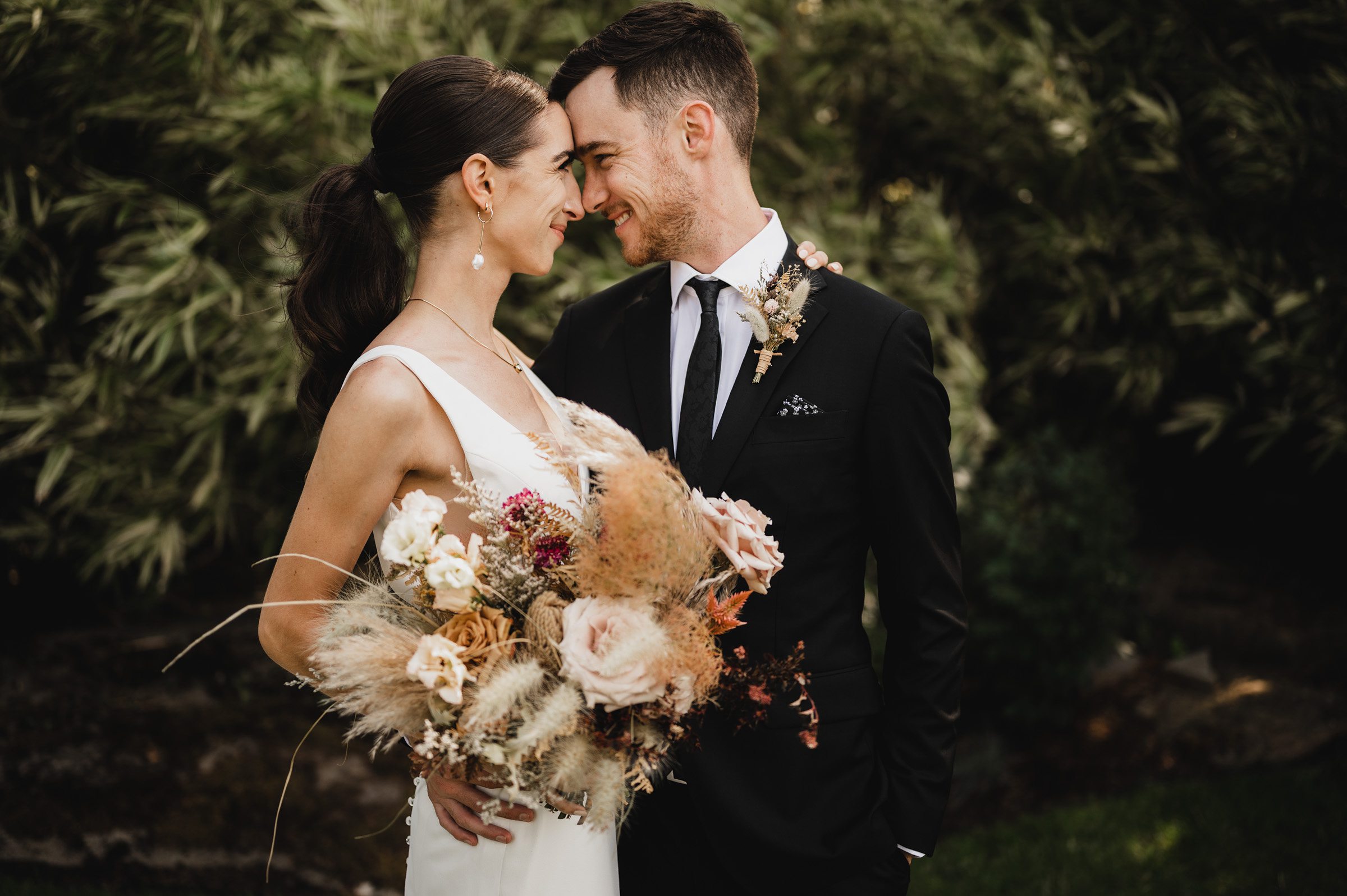 The bride and groom exchange smiles, holding a bouquet outdoors during their wedding photos at The Griffin House.