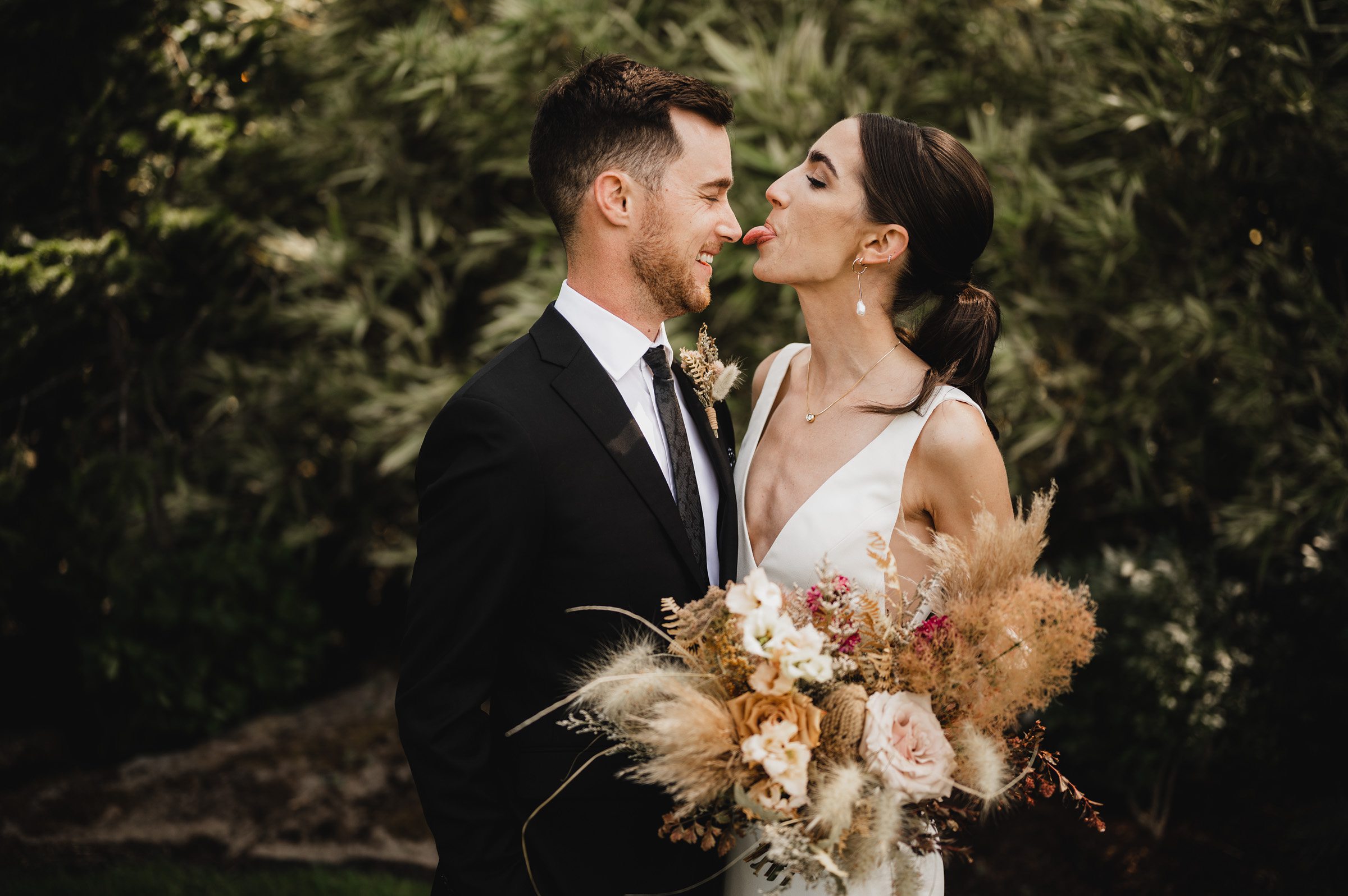 At The Griffin House, a bride playfully sticks her tongue out at the groom while holding a bouquet of dried flowers, set against a backdrop of lush greenery.