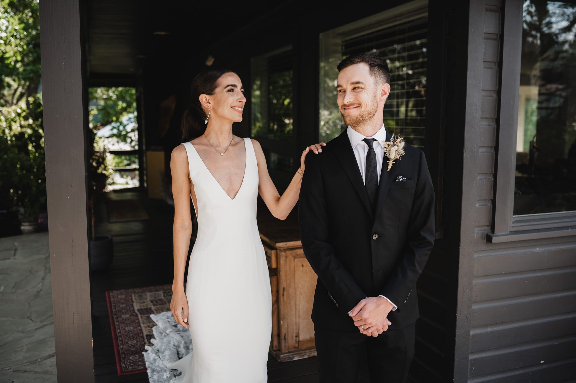 The bride, in a stunning white gown, smiles at her groom in a black suit while standing on the porch. It’s one of those perfect moments captured at The Griffin House during their wedding photo session.