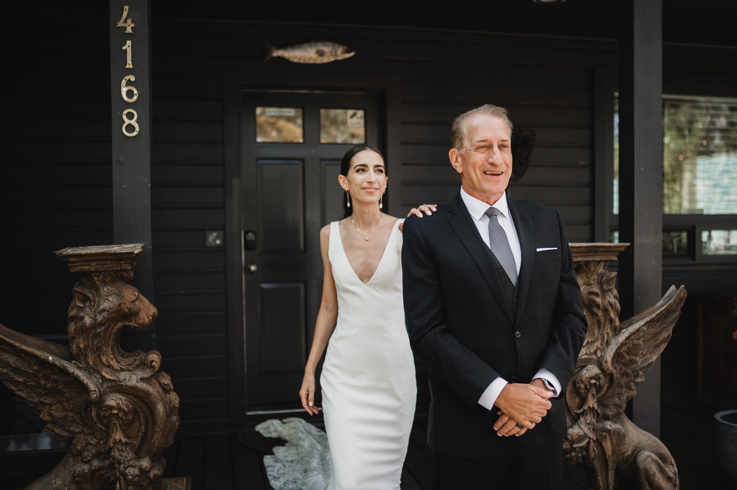 The bride in a white dress rests her hand on the smiling mans shoulder as they stand outside the dark house, numbered 4168. Both are in formal attire, capturing a perfect moment for their wedding photos at The Griffin House.