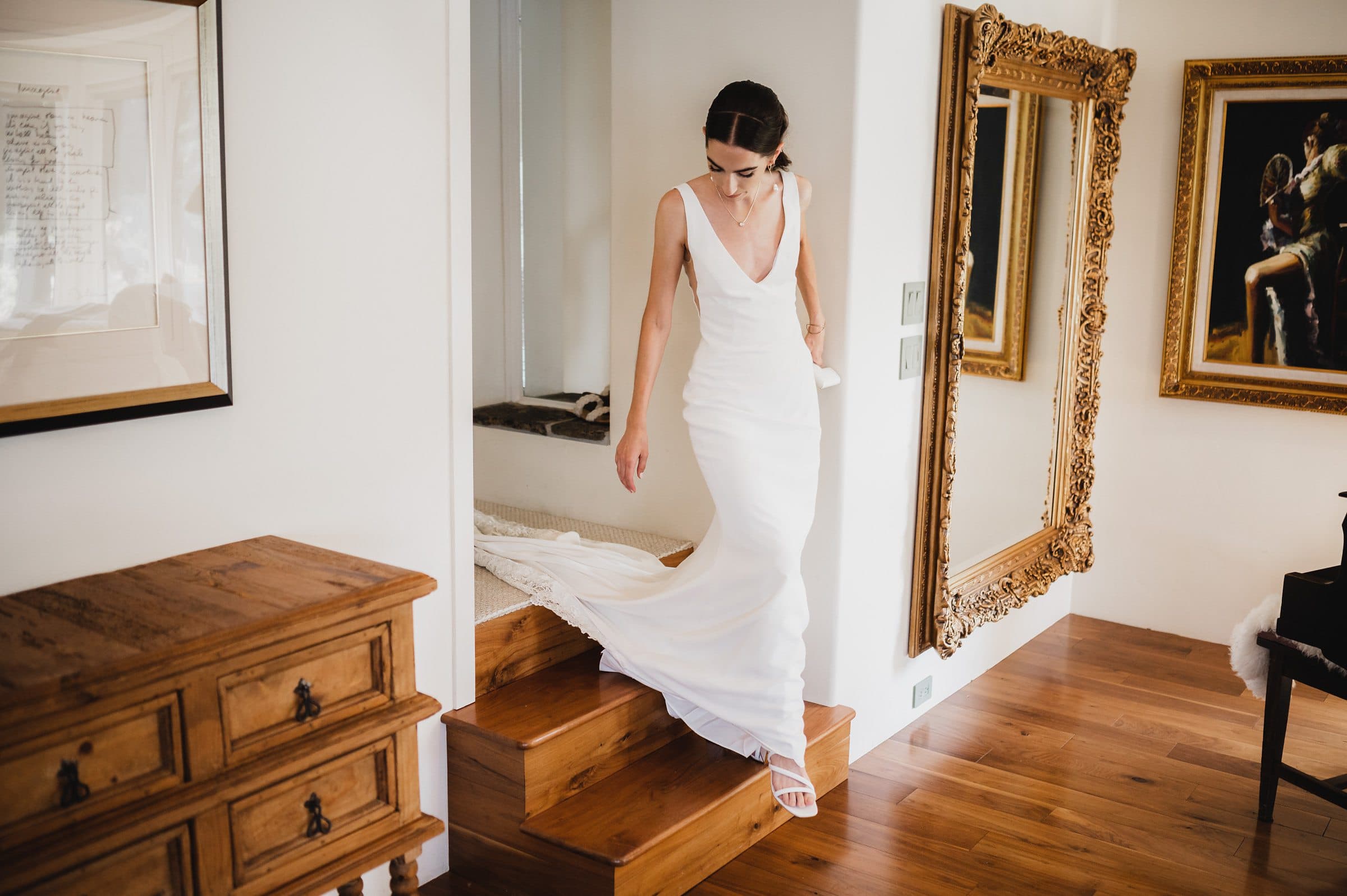 The bride, draped in a white dress, gracefully descends wooden steps in a well-decorated room adorned with mirrors and paintings, capturing perfect wedding photos at The Griffin House.