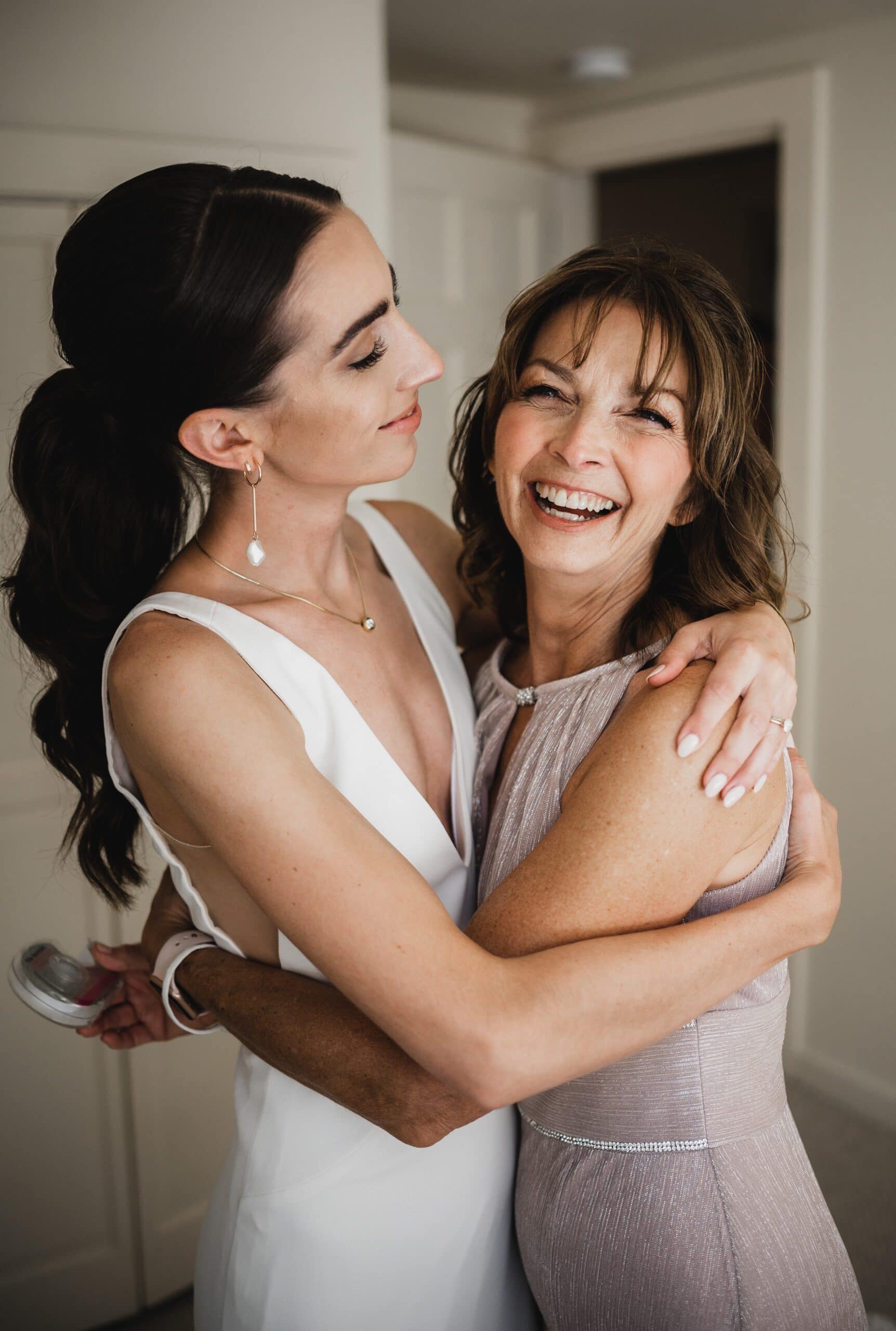The bride in her white gown warmly embraces a her mom who is smiling in a light dress indoors, captured beautifully in wedding photos at The Griffin House.