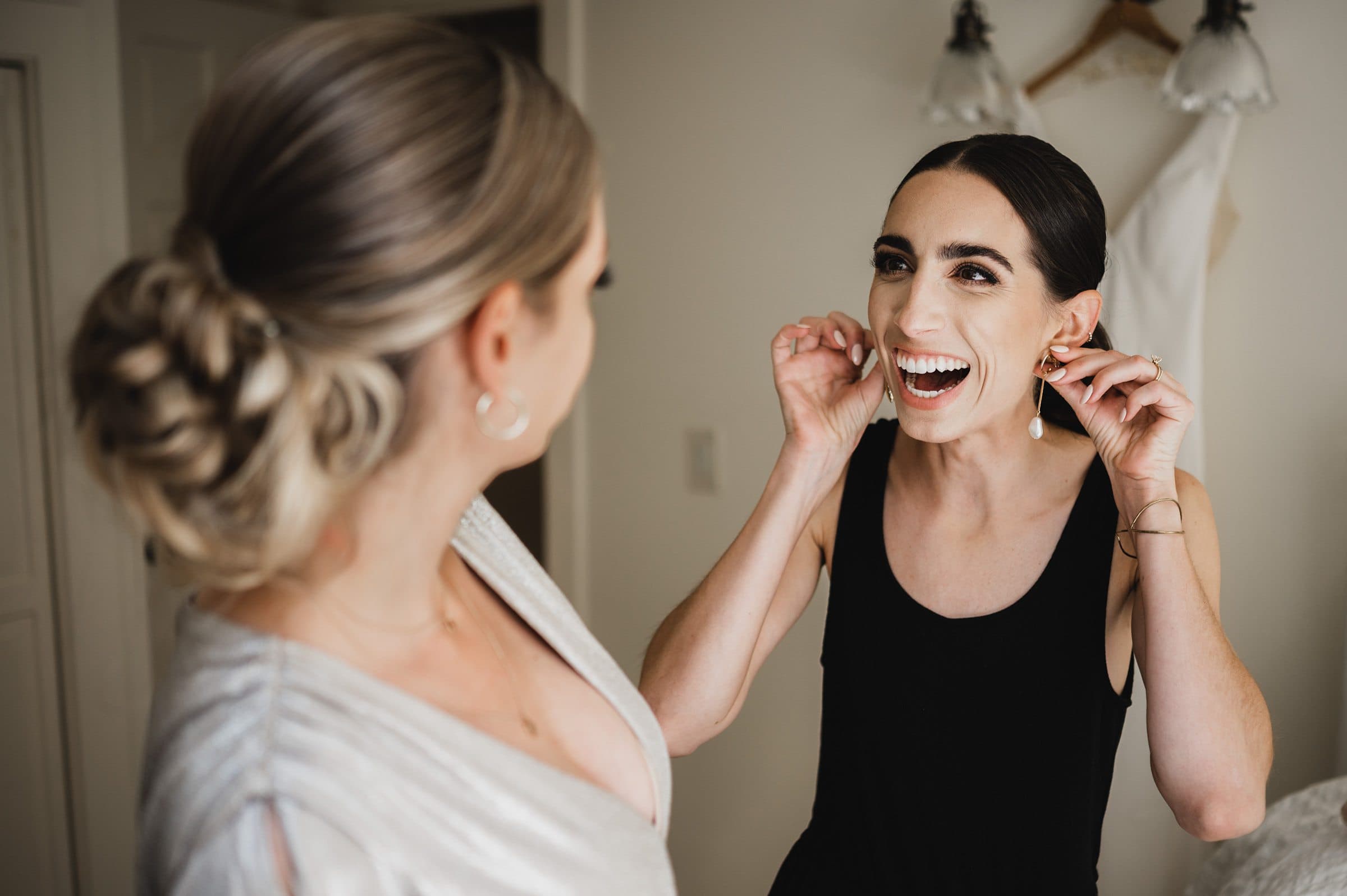 Two women smile warmly as one adjusts her earrings, a beautiful dress hanging in the background. Its a perfect prep moment for wedding photos at The Griffin House.