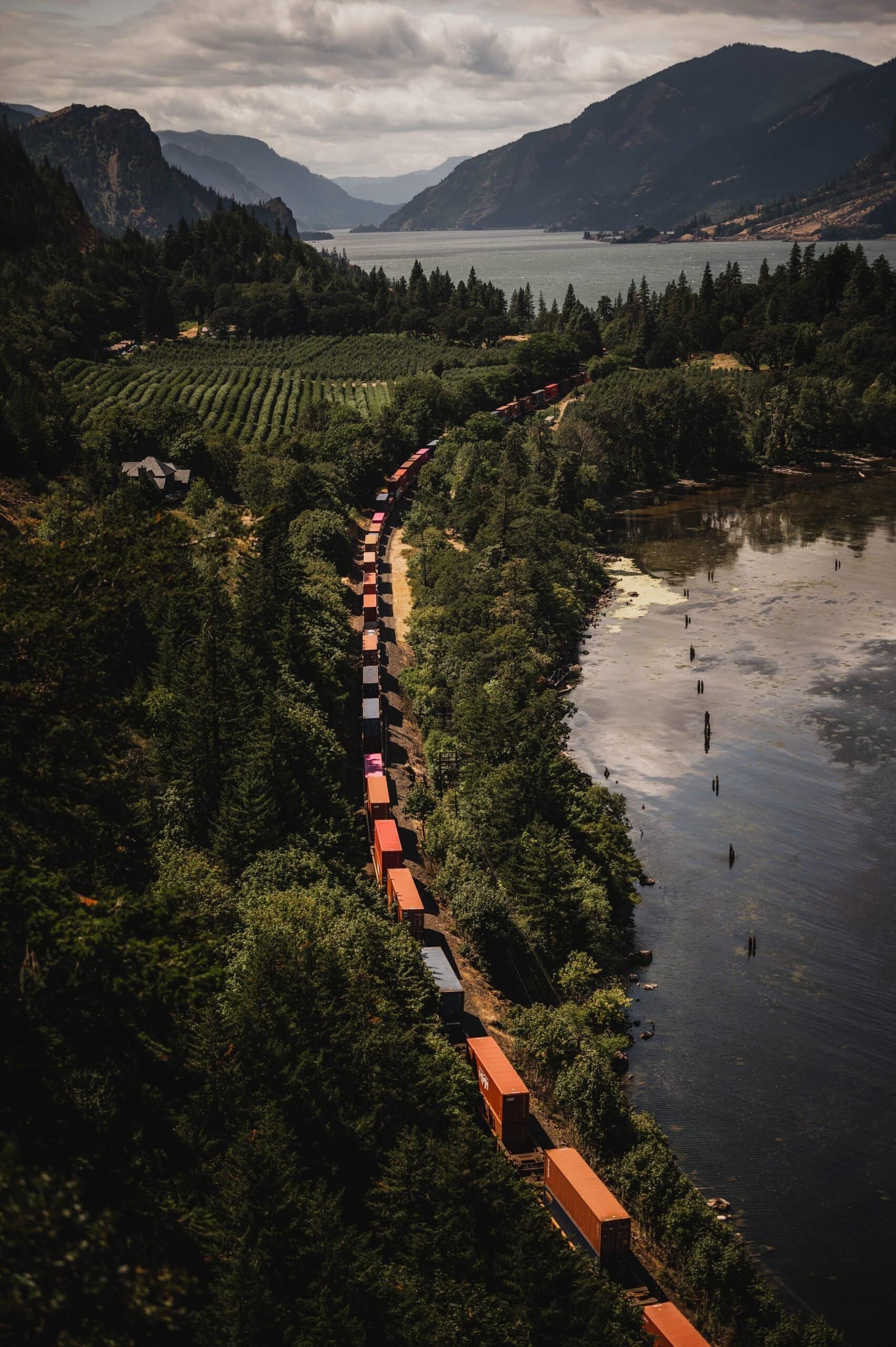 A train with colorful cargo containers winds through a forested landscape by a lake, against the backdrop of majestic mountains, creating a picturesque scene perfect for wedding photos at The Griffin House.