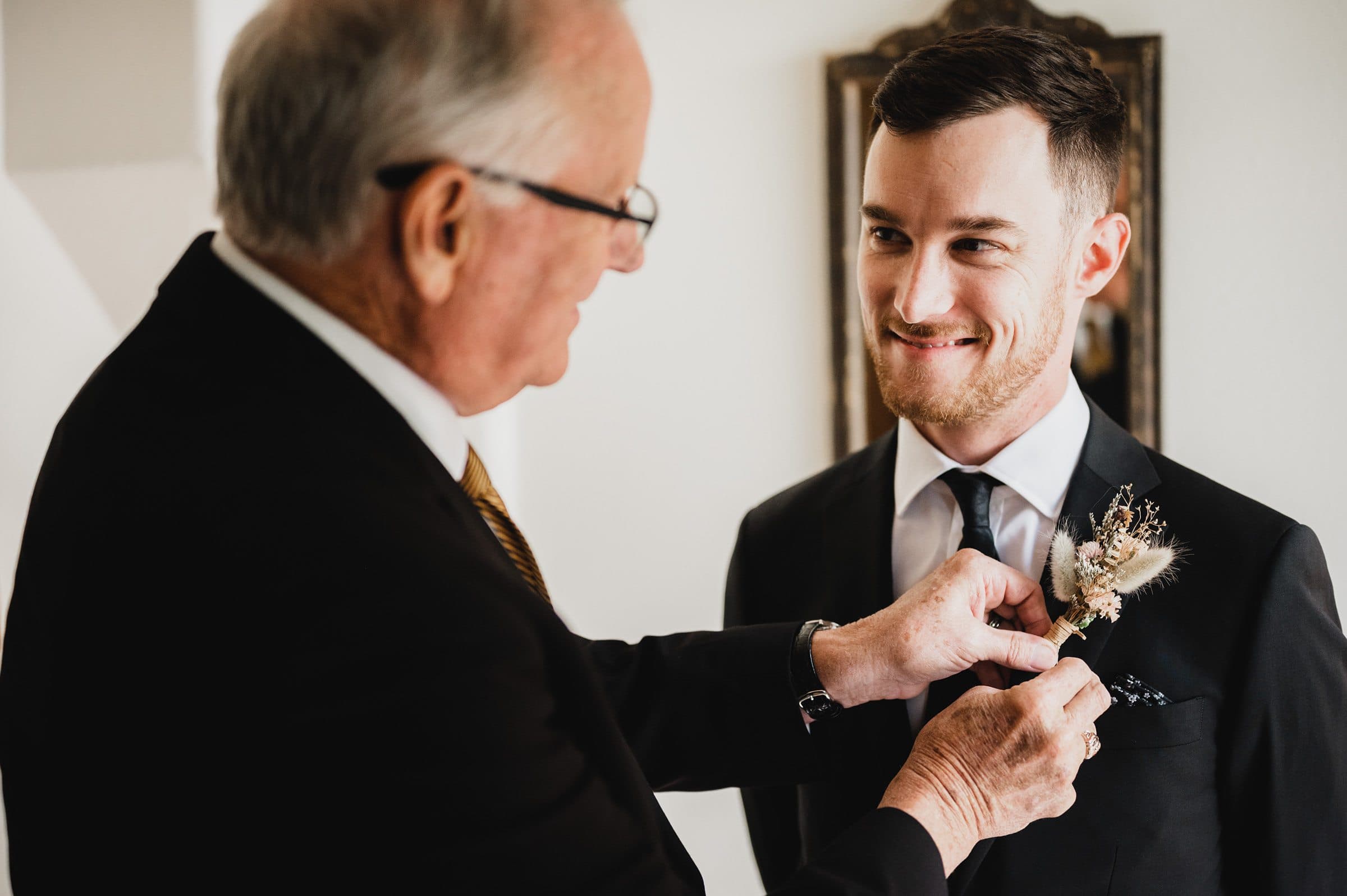At The Griffin House, an older man pins a boutonniere on a smiling groom in a suit, capturing a timeless wedding moment.