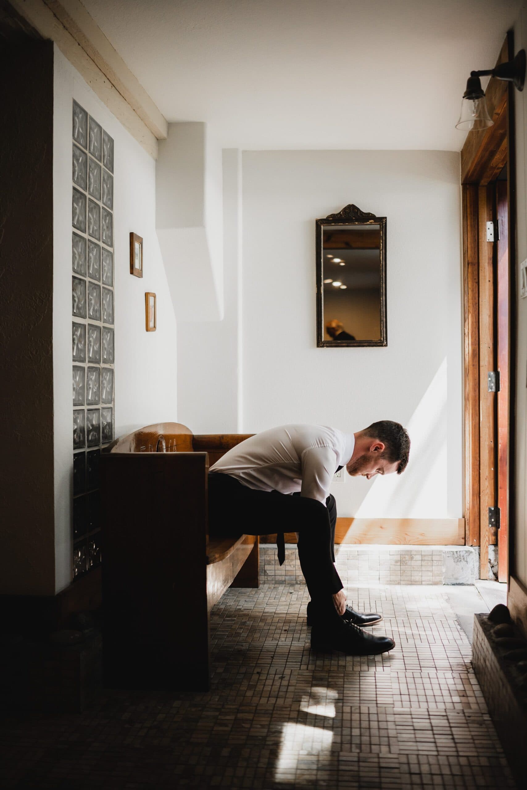 A man sits on a bench in a sunlit hallway, tying his shoe, as light reflects off the mirror on the wall. It’s an intimate moment similar to those captured in wedding photos at The Griffin House.