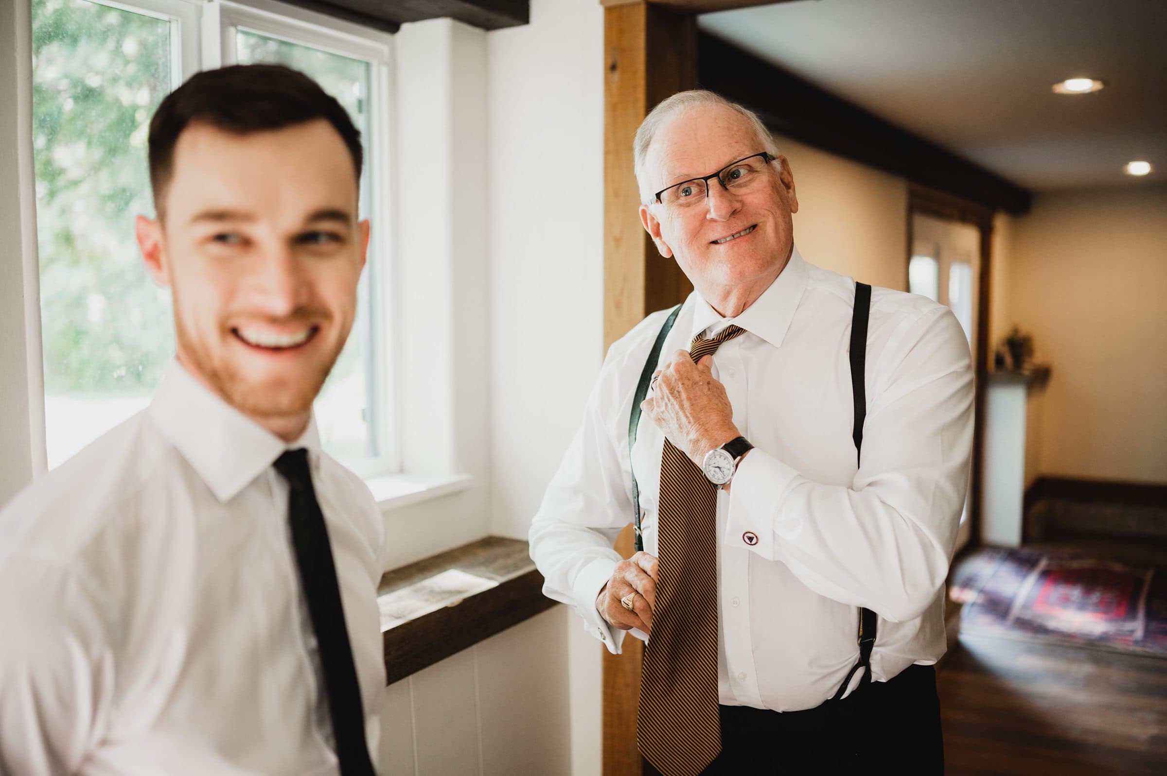 Two men in dress shirts are smiling indoors, one adjusting his tie while the other stands near a window, capturing a candid moment perfect for wedding photos at The Griffin House.