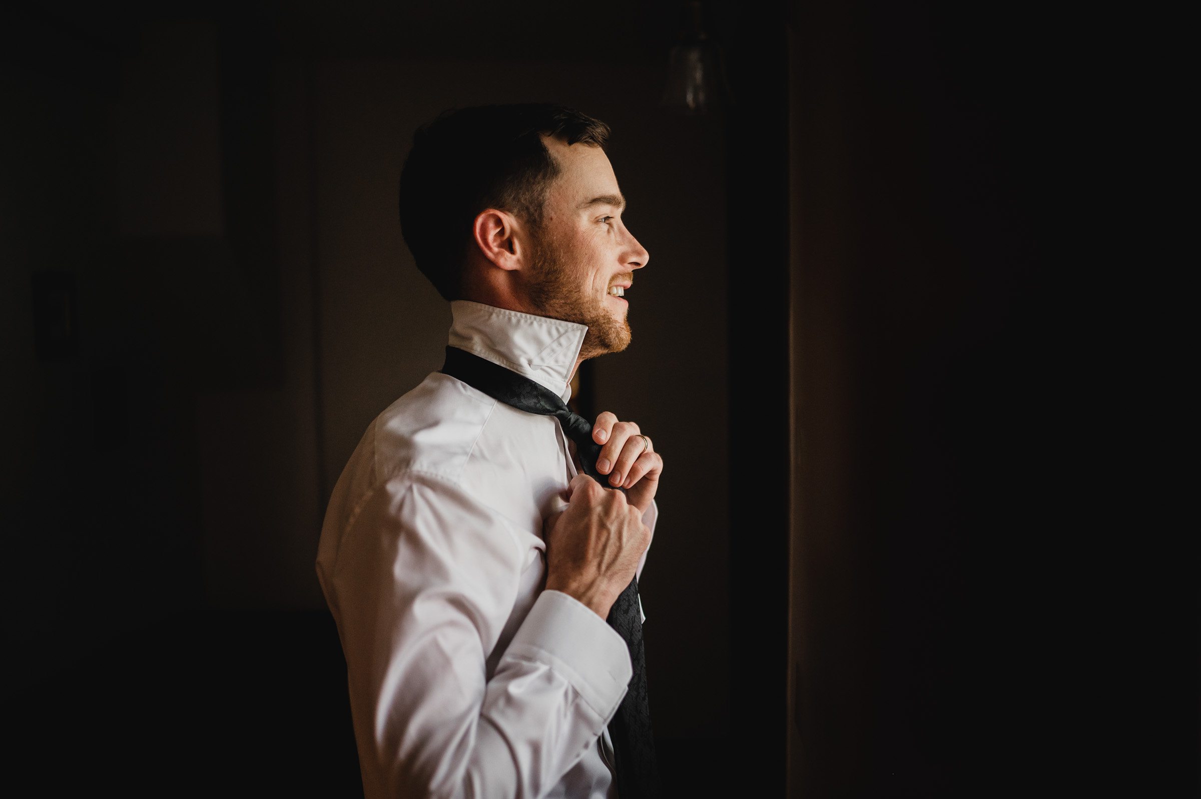 A man in a white shirt smiles while adjusting his black tie near a window, preparing for wedding photos at The Griffin House.