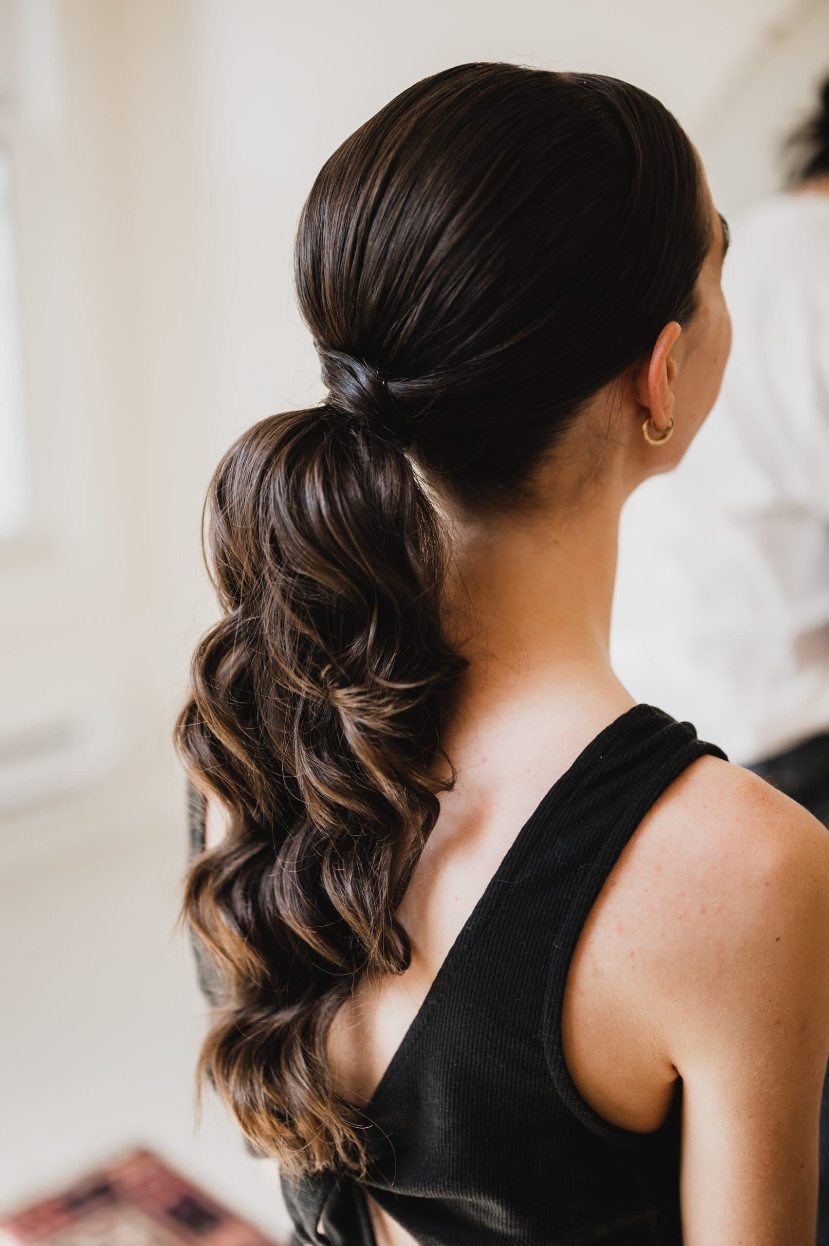 A woman with a long, wavy brown ponytail wearing a black sleeveless top stands in a bright room, perhaps preparing for wedding photos at The Griffin House.