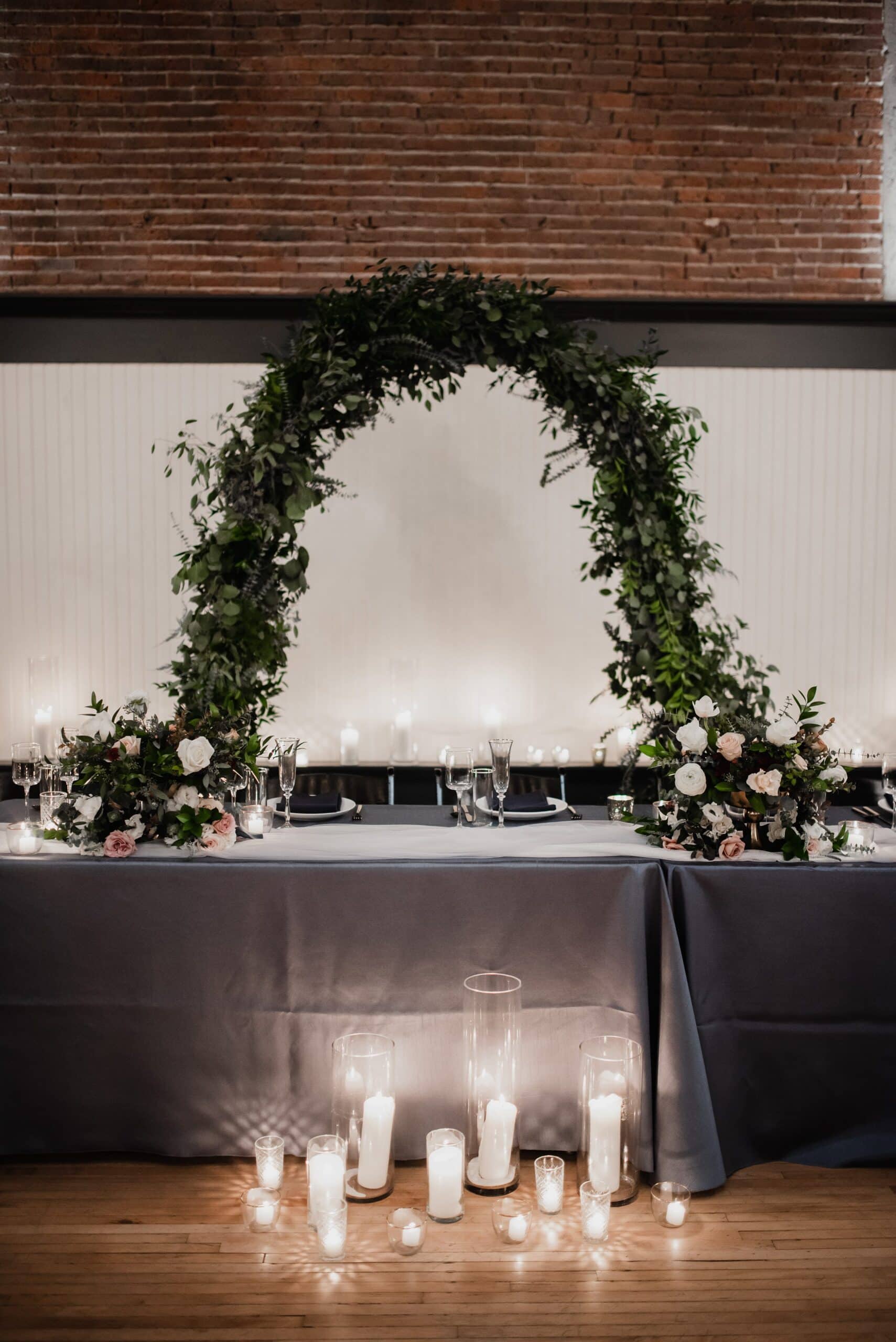 The Evergreen, a Portland wedding venue, features a stunning reception table adorned with a green arch, soft candlelight, and exquisite floral arrangements on its elegant wooden floor.