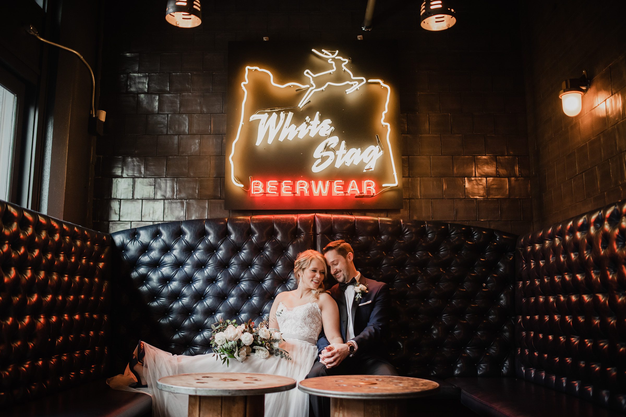 A couple cuddles in a cozy booth at Loyal Legion during their wedding at The Evergreen, under a neon White Stag Beerwear sign, smiling and holding hands.