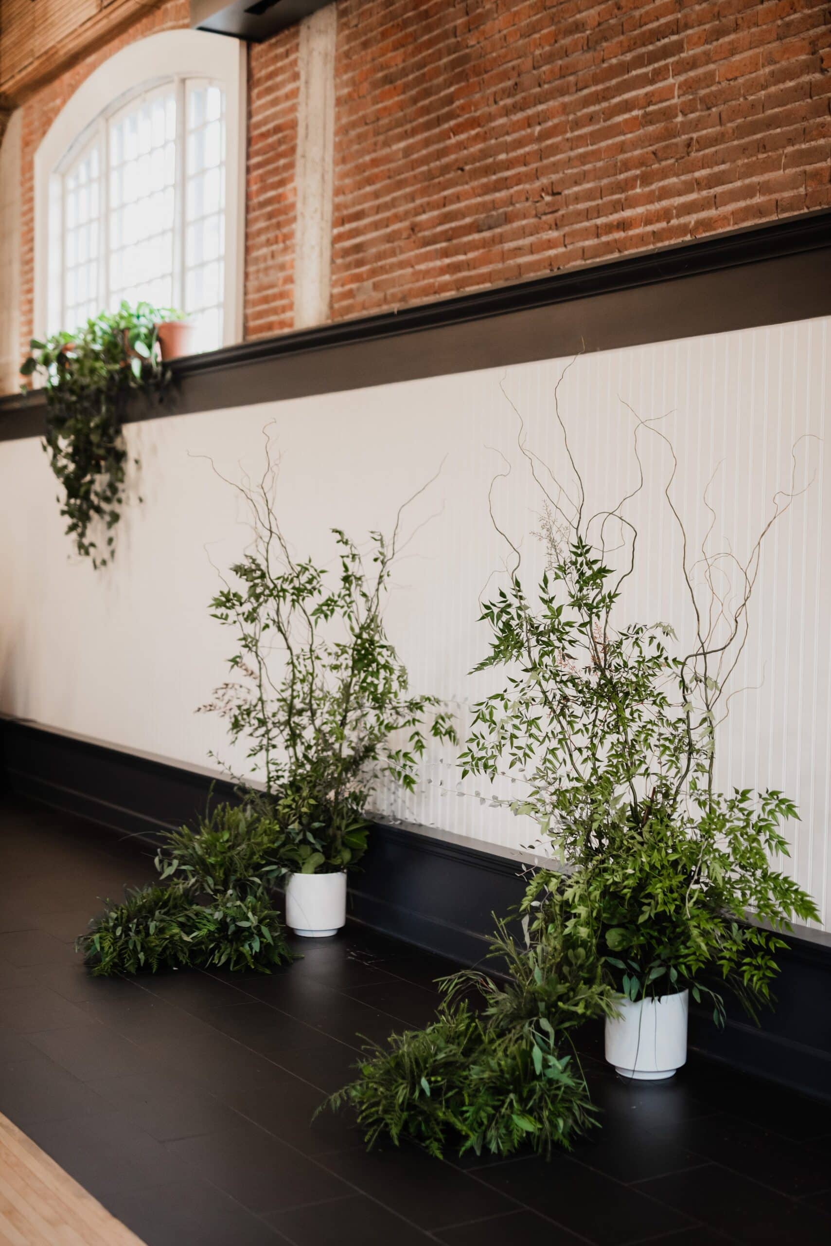 Plants and branches in white pots decorate The Evergreen, a renowned Portland wedding venue, enhancing its brick-walled room with high windows and dark floor.