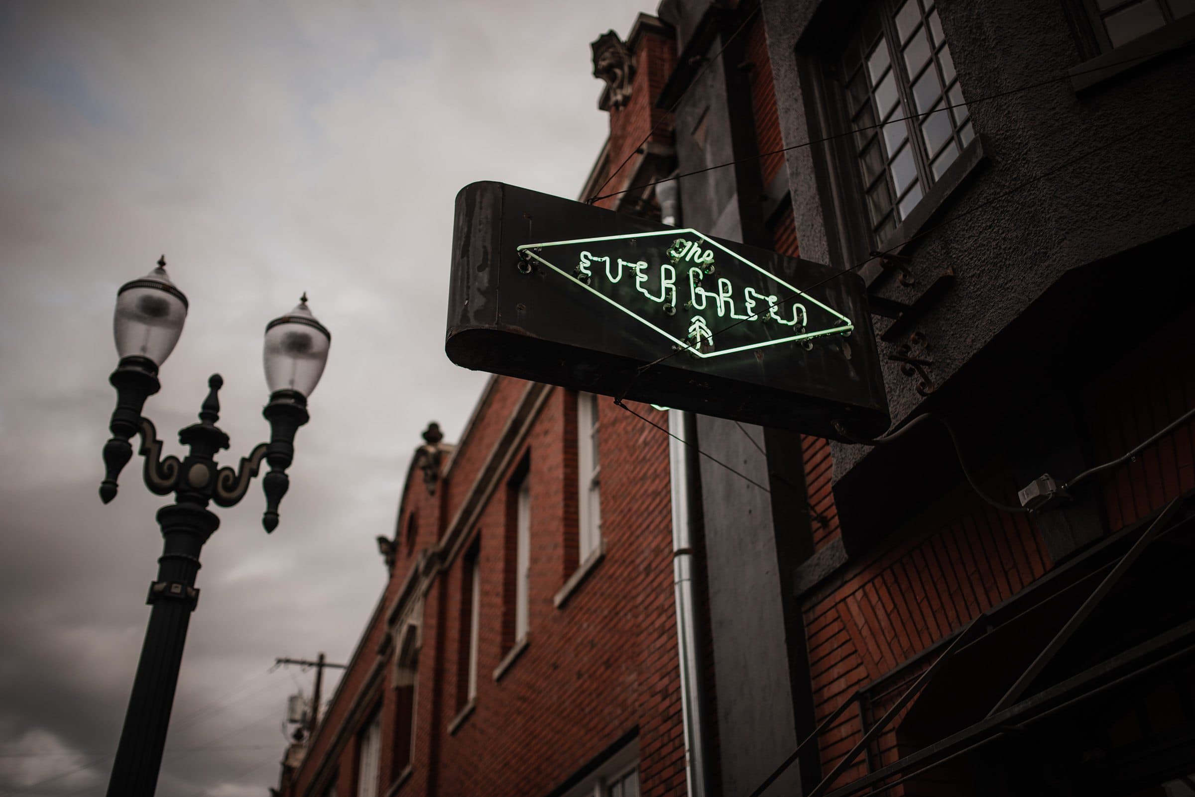A vintage streetlamp casts a nostalgic glow on the neon sign reading The Evergreen, a charming Portland wedding venue nestled against a brick building under a cloudy sky.