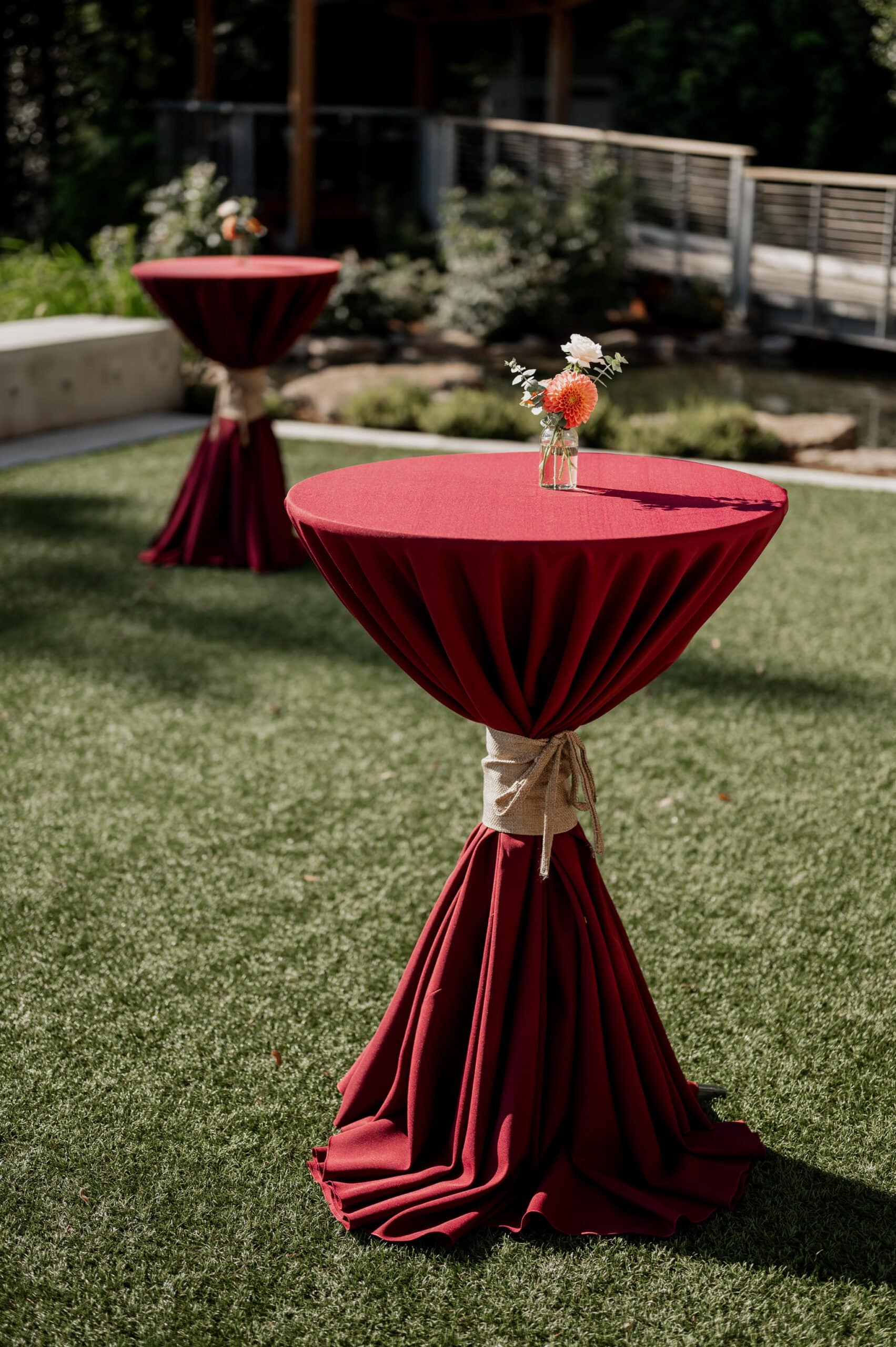 Two tall tables draped in burgundy cloth, each adorned with small flower vases, stand gracefully on the lush grounds of Amaterra Winery.
