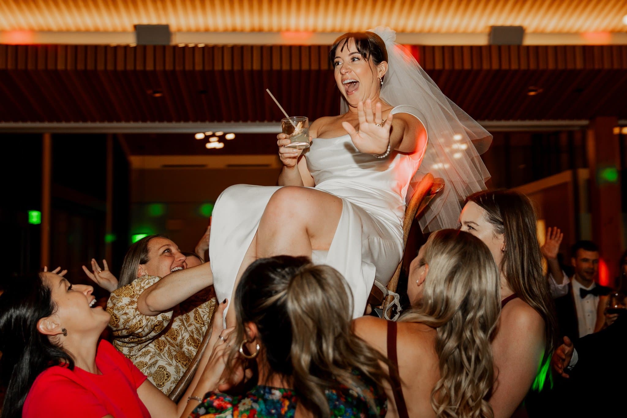 At the Skyline Ballroom at Amaterra Winery, a popular Portland wedding venue, the bride in her white dress and veil is joyfully lifted by friends during the lively celebration, holding a drink and smiling.