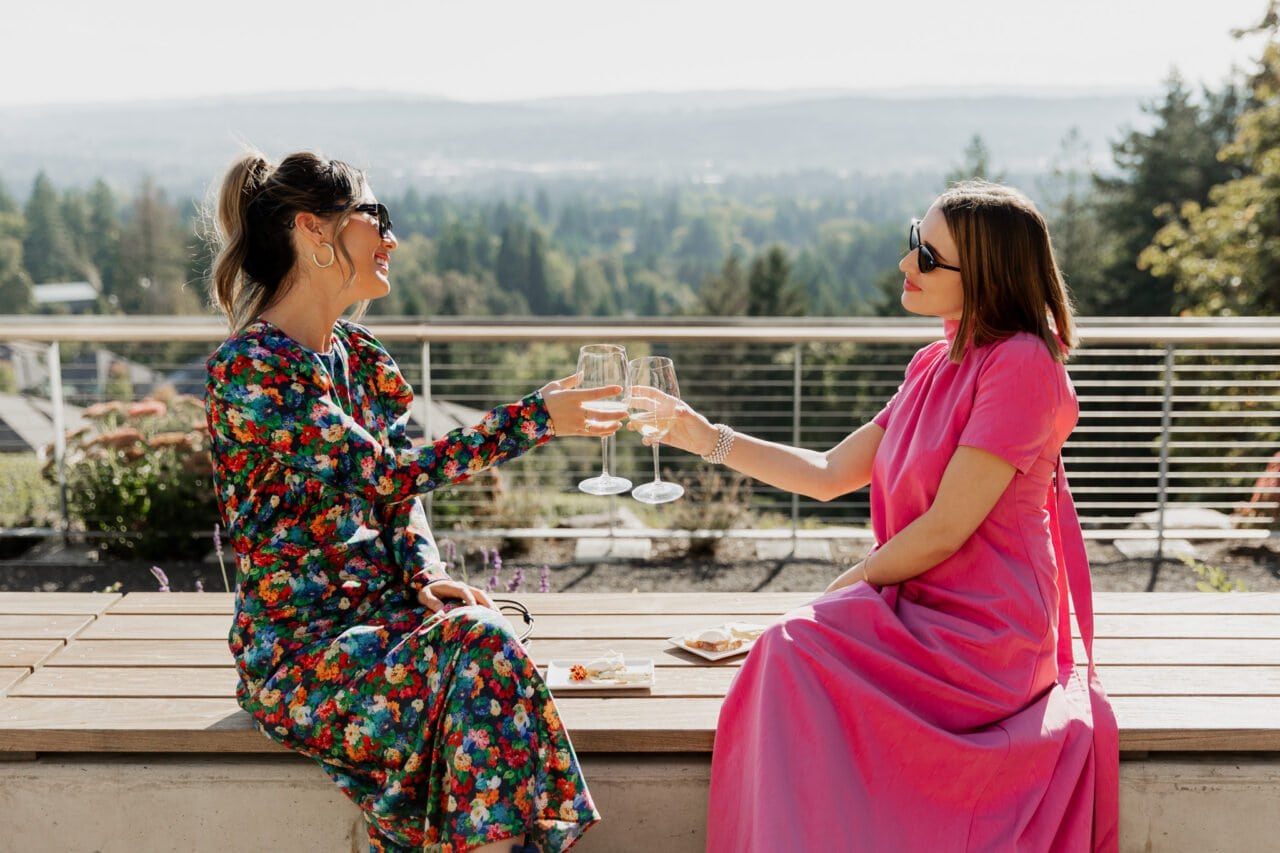 Two women, one in a floral dress and the other in pink, sit outdoors clinking wine glasses. The scene captures the joyful essence of Amaterra Winery wedding photos.