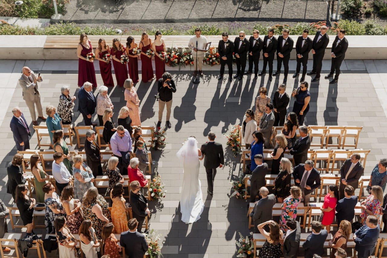 Aerial view of an outdoor Amaterra Winery wedding, with the bride and groom gracefully walking down the aisle, as guests seated on both sides capture photos of the enchanting moment.