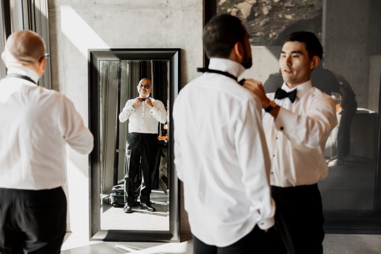 Three men in formal attire adjust bow ties; one is seen in a mirror reflection. This elegant moment is beautifully captured among the vineyards, perfect for any Amaterra Winery wedding photos collection.
