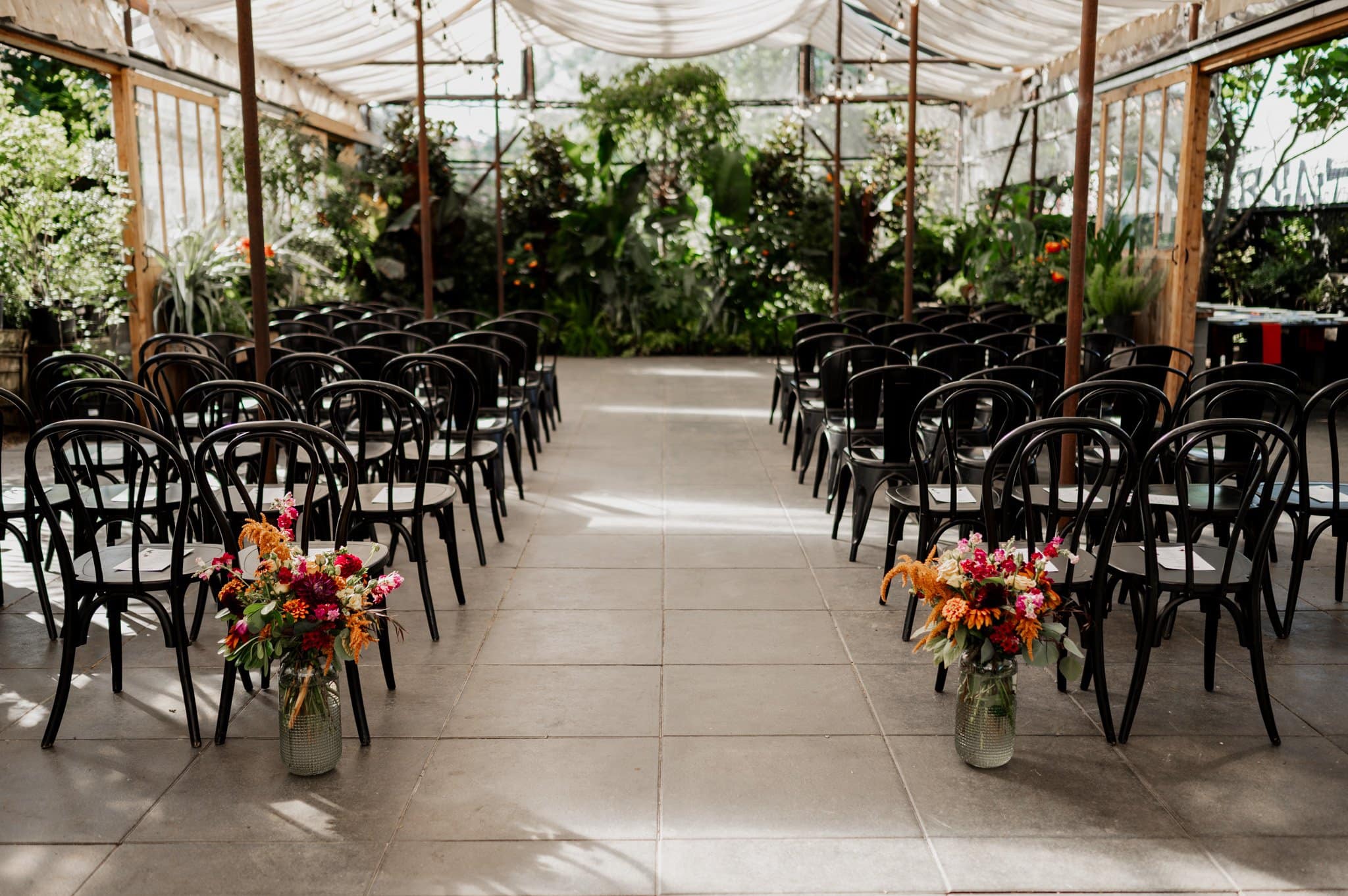 An aisle with black chairs and colorful floral arrangements leads to a garden backdrop under a translucent canopy at Block House, a charming Portland wedding venue.