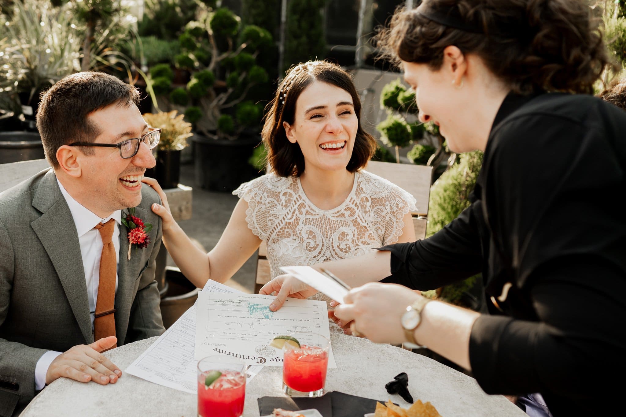 A smiling couple, holding a document, shares a joyful moment with a woman at Blockhouse, a renowned Portland wedding venue, surrounded by drinks and lush plants.