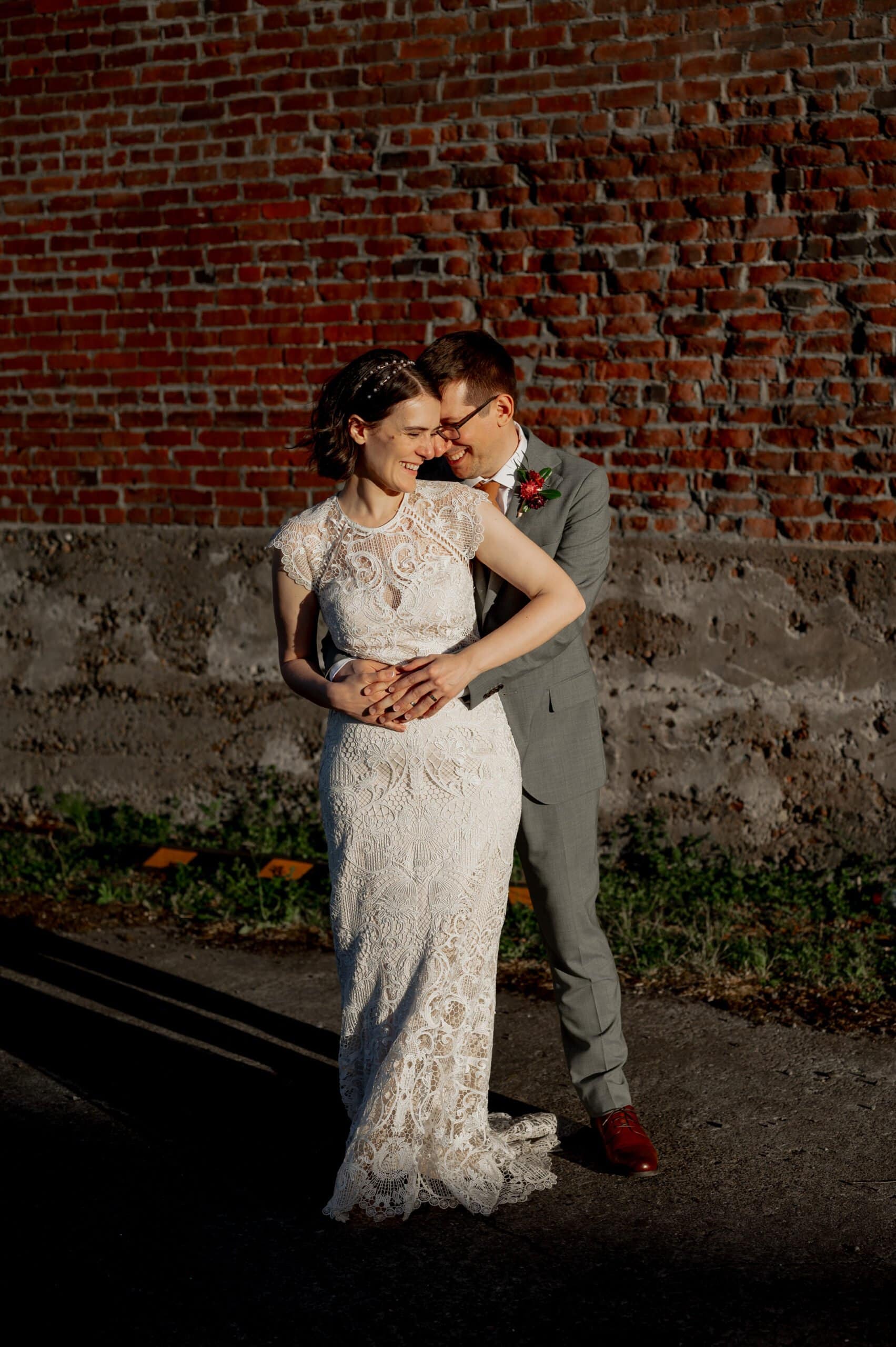 A couple in wedding attire embraces in front of a brick wall, sunlight highlighting their faces and clothes, near Blockhouse, a Portland wedding venue.