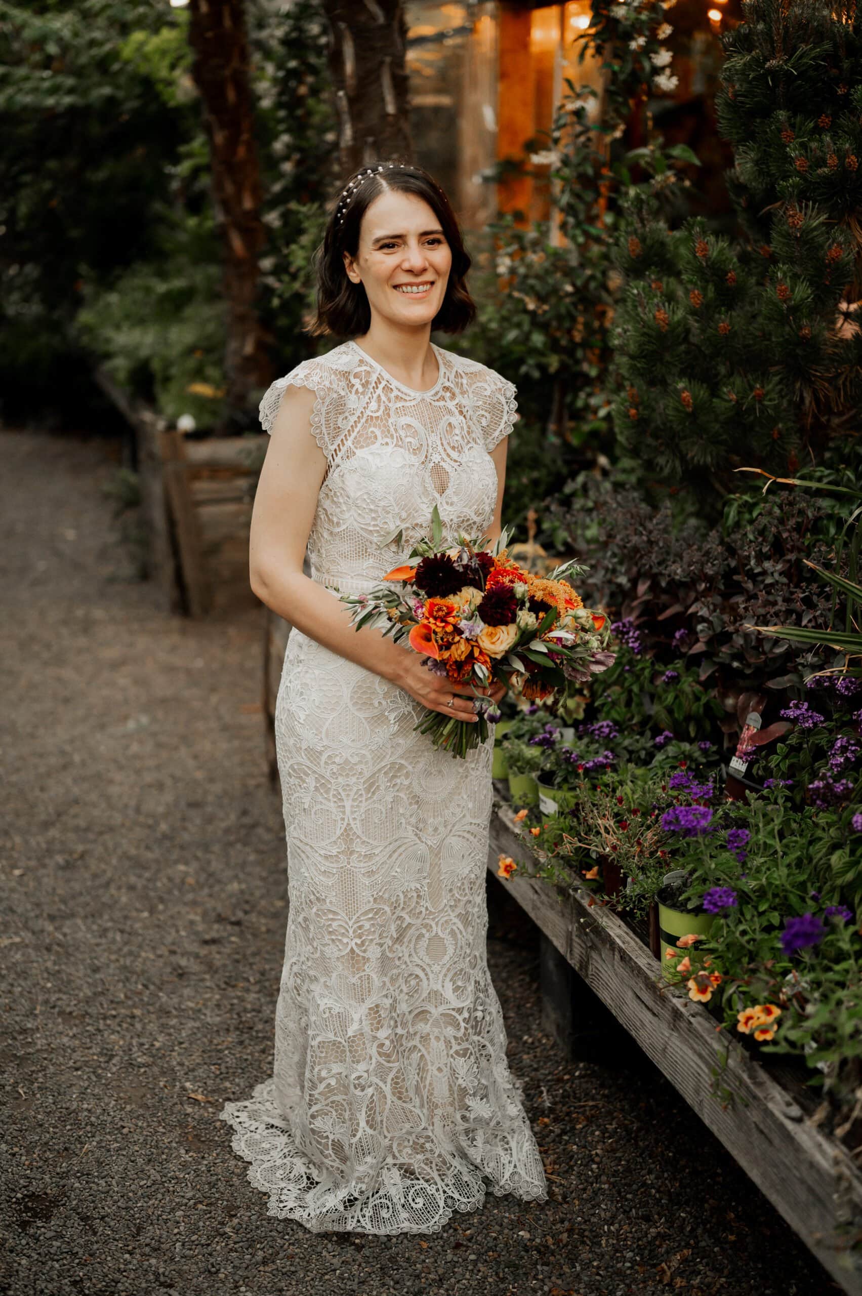 Bride in a lace wedding dress holds a colorful bouquet, standing beside a garden bed with purple flowers at Blockhouse, a Portland wedding venue.