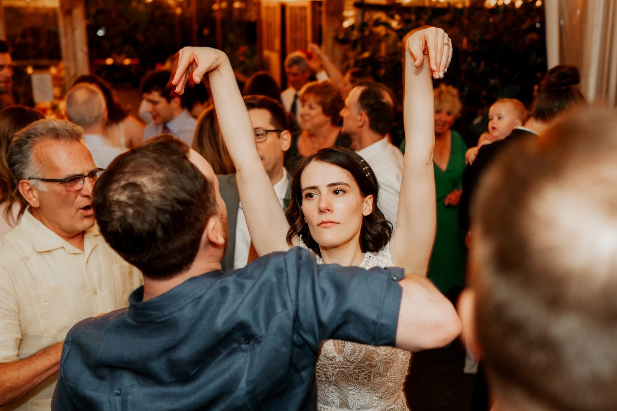 A woman dances with a partner at a lively indoor gathering in Blockhouse, where people mingle and a baby is held in the background, creating the perfect setting for wedding photos.