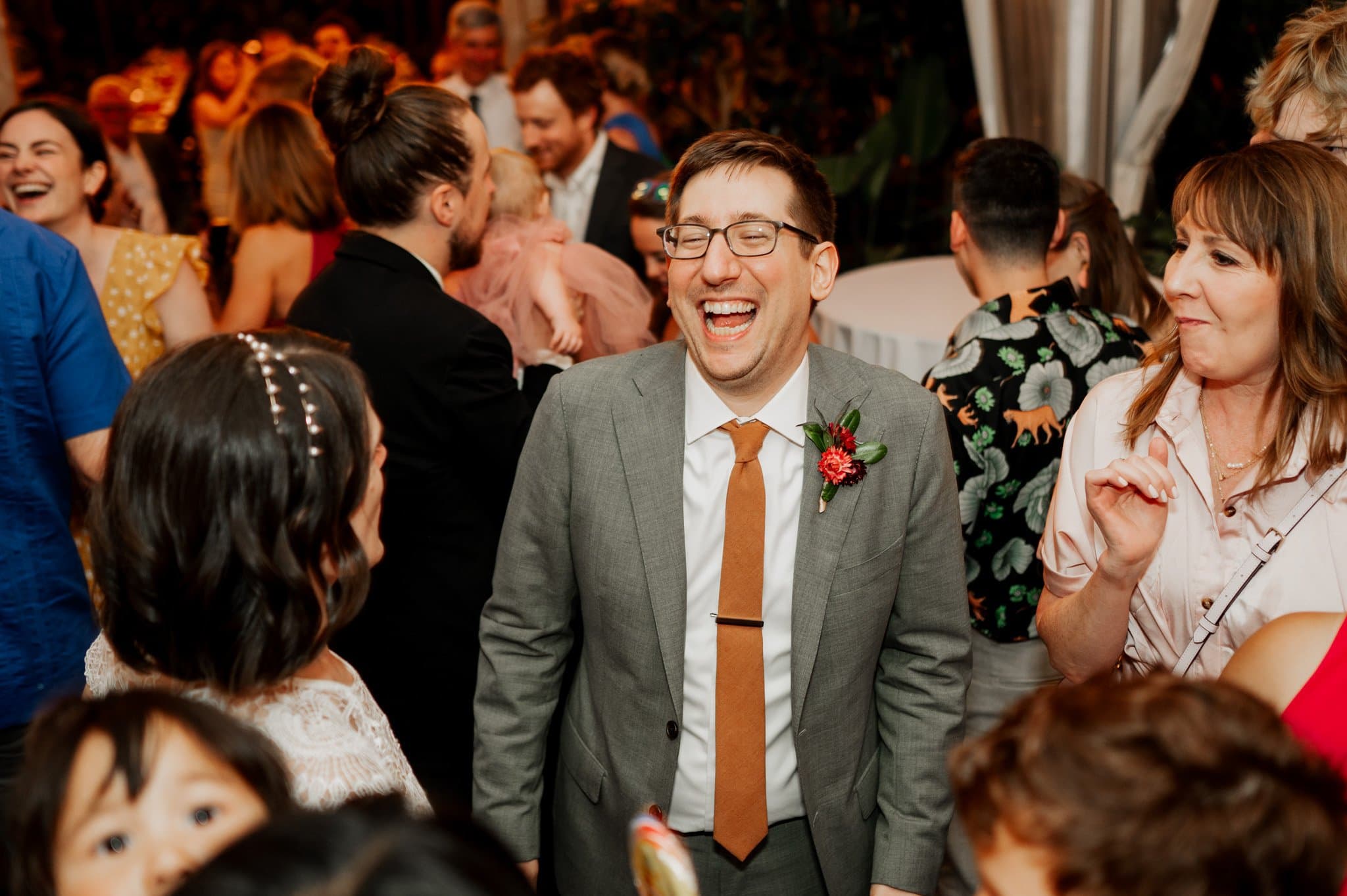 A man in a suit laughs joyfully at a festive gathering, surrounded by people chatting and smiling. The lively scene is captured like wedding photos at Blockhouse, with everyone enjoying the cheerful atmosphere.