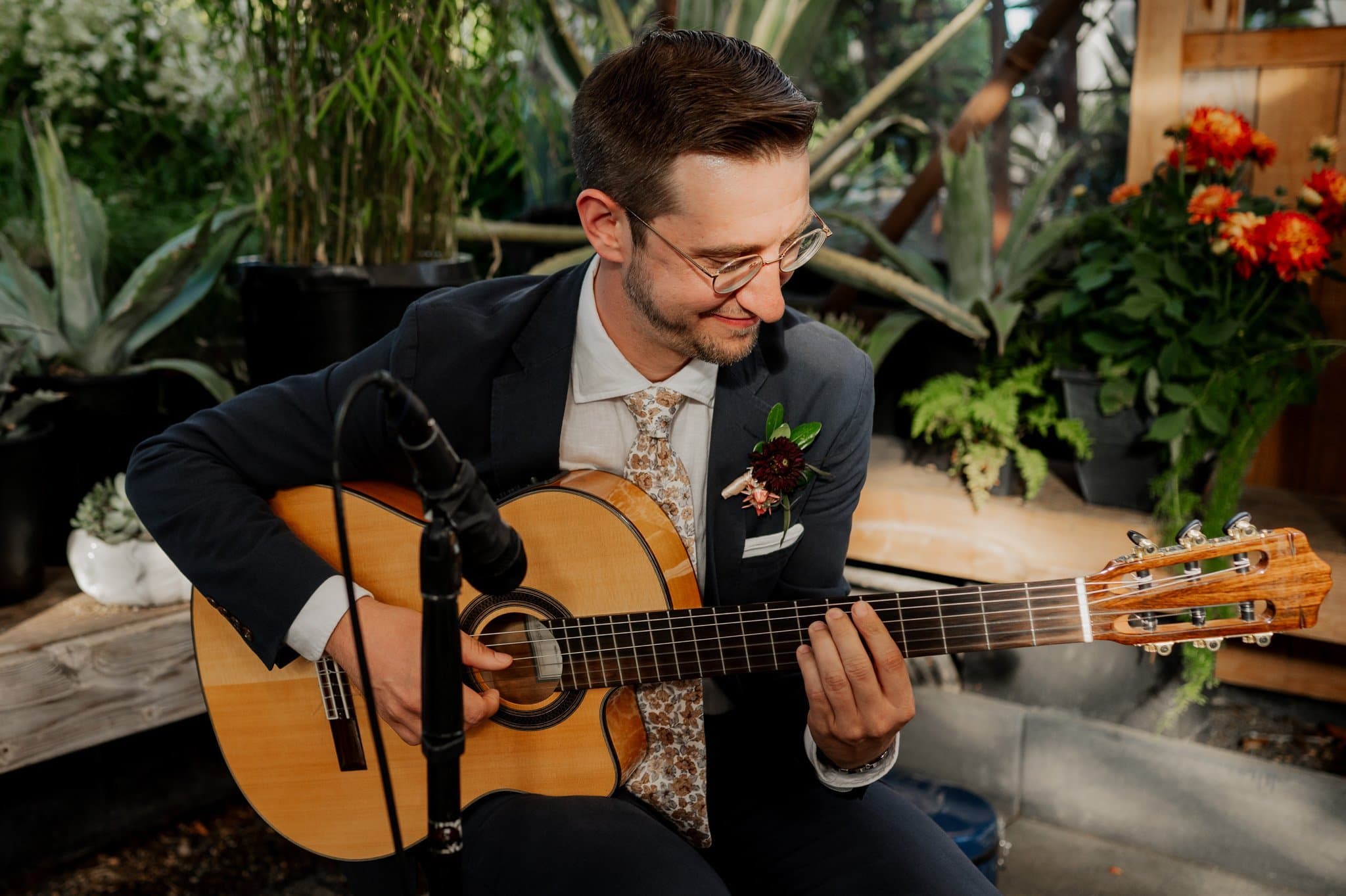 A man in a suit strums a guitar, surrounded by lush plants and vibrant orange flowers. The idyllic garden setting evokes the charm of wedding photos at Blockhouse, creating an atmosphere filled with romance and elegance.