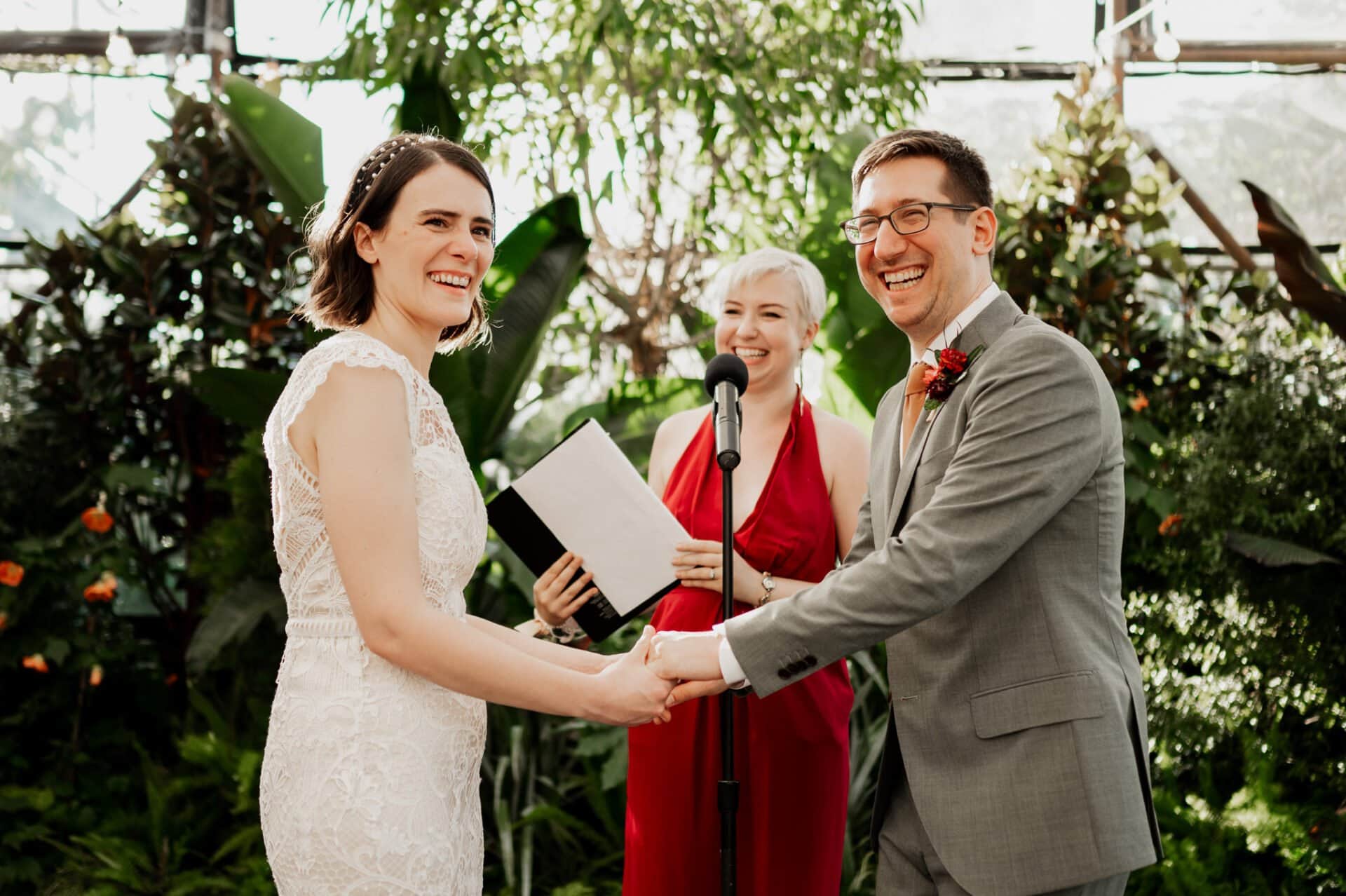A couple holding hands and smiling during a wedding ceremony at Blockhouse, surrounded by a lush, green environment.