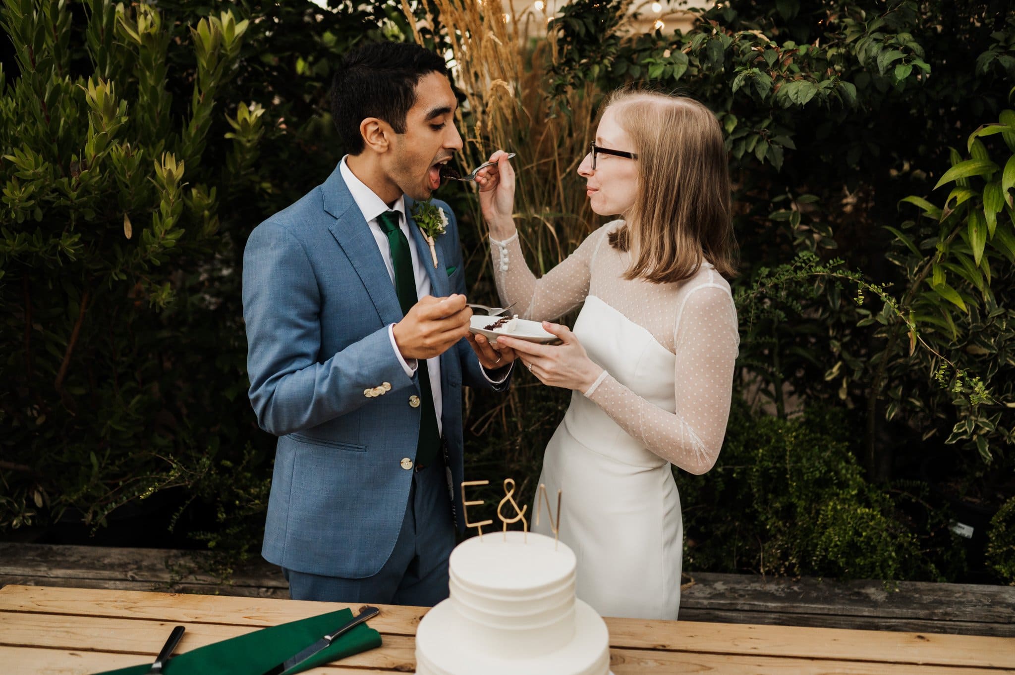 A couple stands outdoors, feeding each other cake, captured beautifully in Blockhouse wedding photos. The man is in a blue suit, the woman in a white dress. A white cake with an E & N topper graces the table before them.