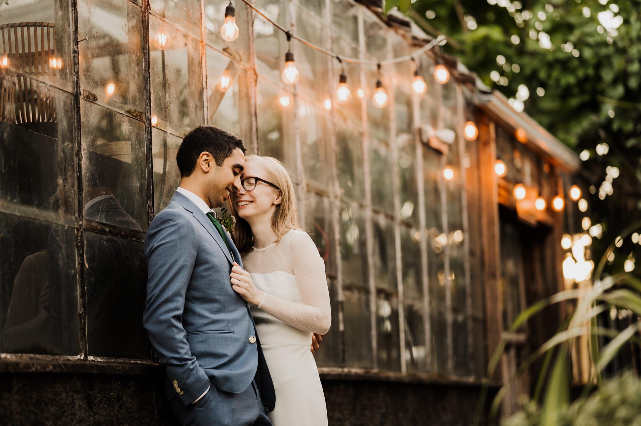 A couple stands close, smiling near a glass building adorned with string lights. The man dons a blue suit, and the woman is in a white dress with glasses—perfect moments captured as if from Blockhouse wedding photos.