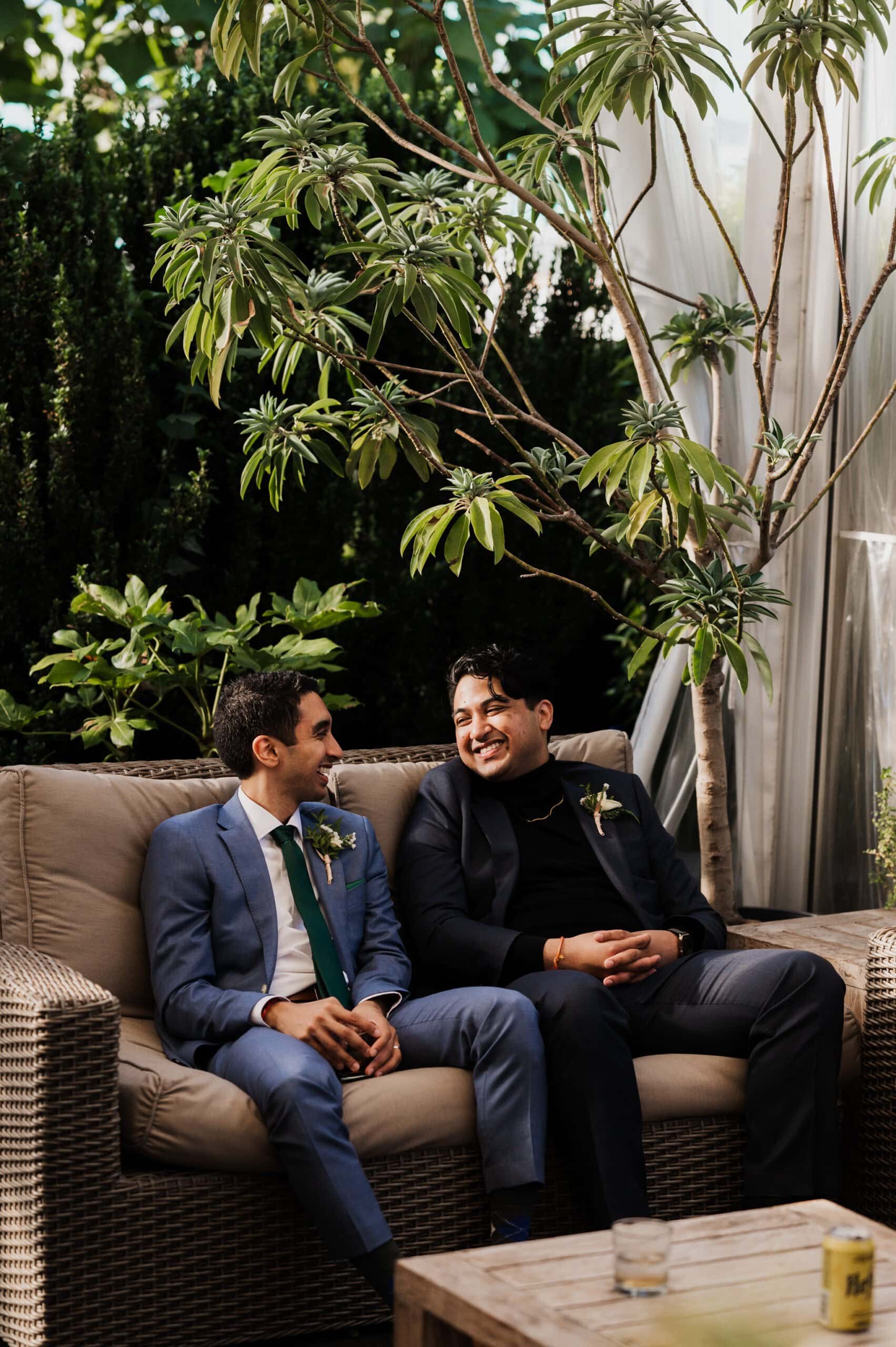 Two men in suits sit on a wicker sofa outdoors, smiling at each other amidst lush greenery, reminiscent of Blockhouse wedding photos.