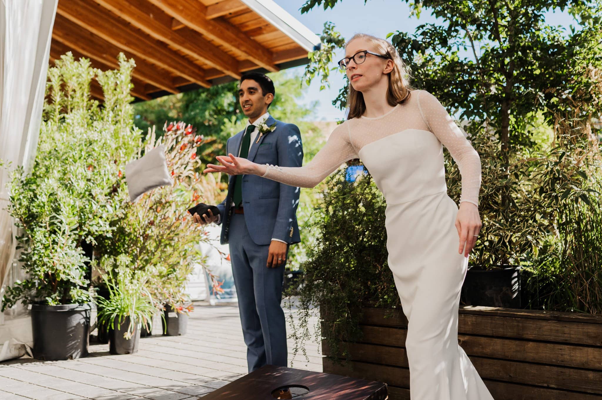 A woman in a white dress tosses a bean bag while a man in a suit watches intently. Captured outdoors amid lush greenery and the rustic charm of the wooden structure, this scene embodies the joyful essence of Blockhouse wedding photos.