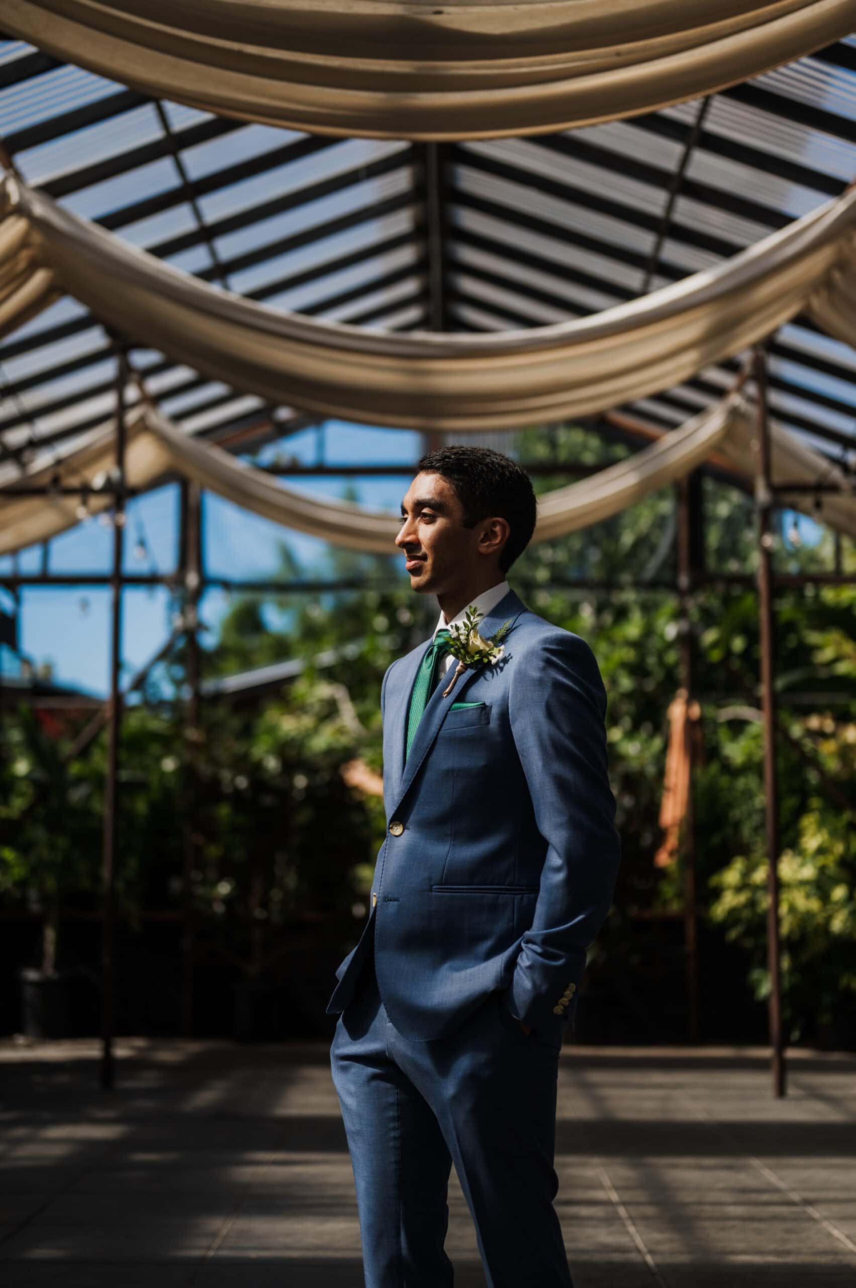 A groom in a blue suit stands indoors under a draped fabric ceiling, surrounded by lush greenery and natural light, capturing the essence of Blockhouse wedding photos.