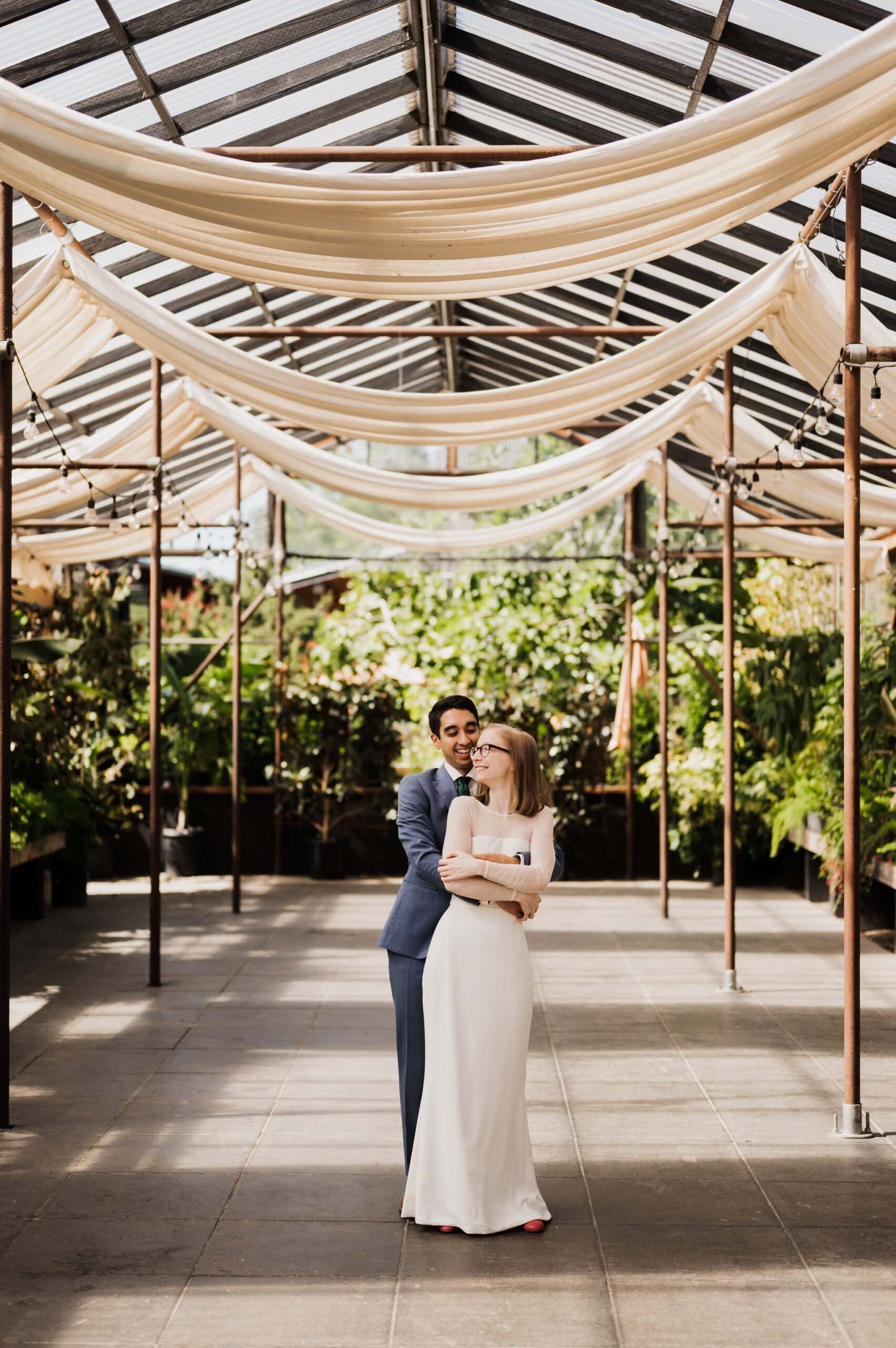 In an enchanting greenhouse, reminiscent of a Blockhouse wedding, a couple embraces amidst the draped fabric and lush greenery in the background of a greenhouse.