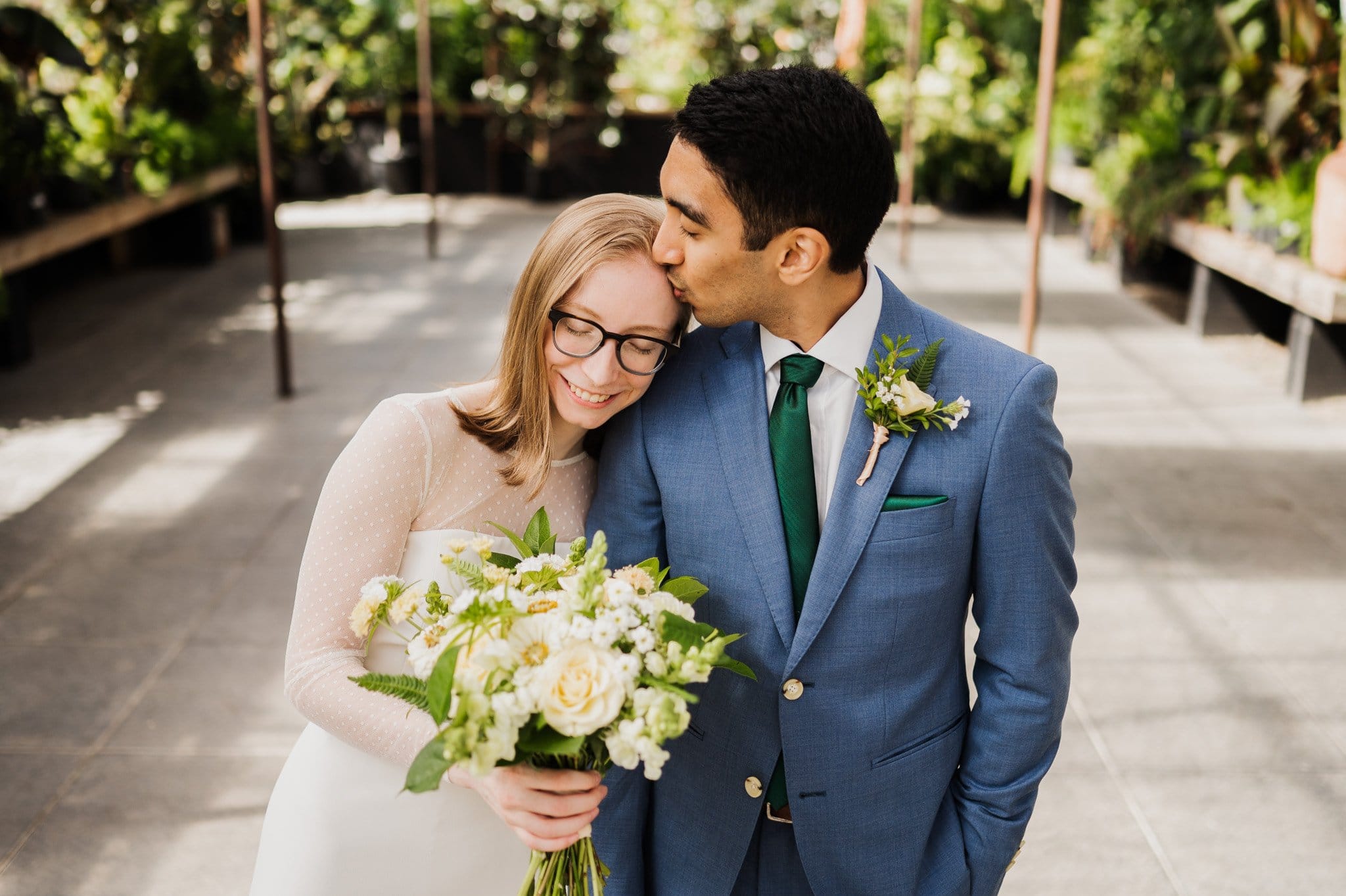 Bride in a white dress holding a bouquet, smiling with eyes closed. Groom in a blue suit kisses her on the forehead amidst lush greenery, capturing the essence of Blockhouse wedding photos.