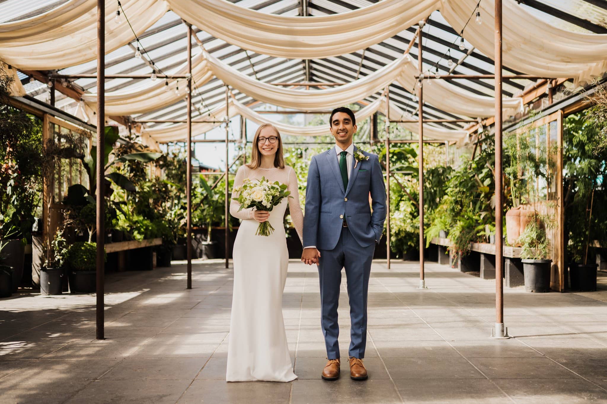 A couple poses indoors in a greenhouse for enchanting blockhouse wedding photos, holding hands. The woman is in a white dress with a bouquet, and the man is in a blue suit. Plants and draped fabric decorate the setting.