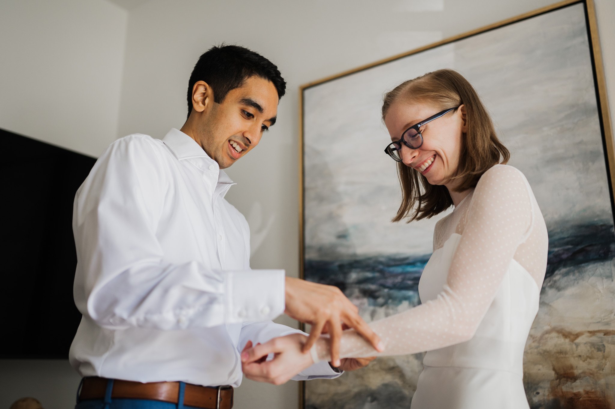 A smiling couple stands indoors, with the man adjusting the womans sleeve near abstract art on the wall, capturing a candid moment reminiscent of elegant Blockhouse wedding photos.