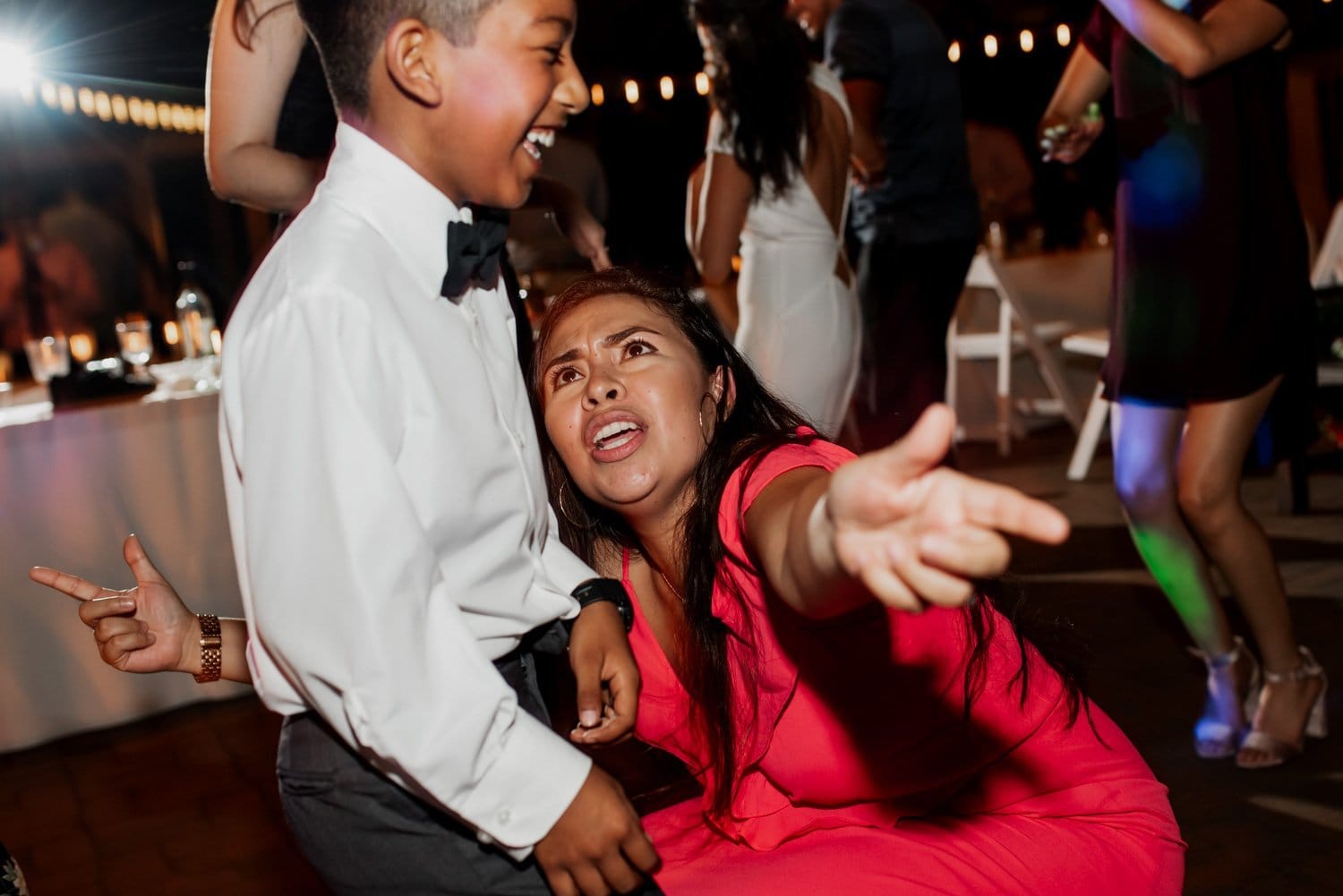 At a Leach Botanical Garden wedding, a woman wearing a red dress gestures enthusiastically as she interacts with others, while a young boy in a bow tie stands nearby. The scene is filled with lively celebration and greenery in the background.