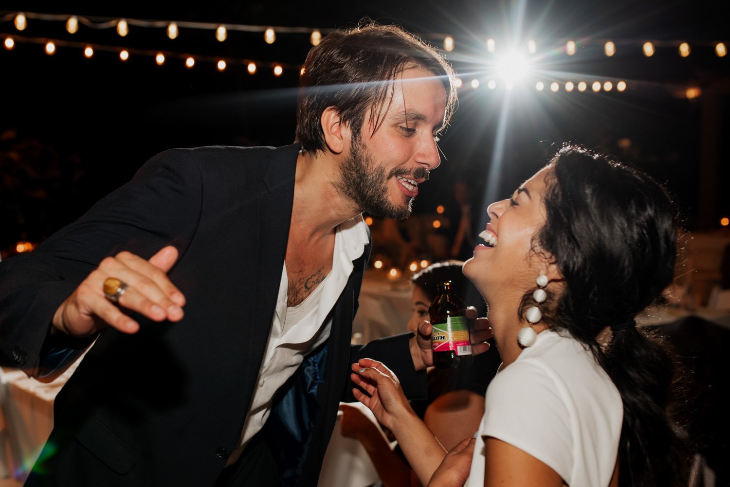A man and woman laugh joyfully under string lights at a nighttime event.